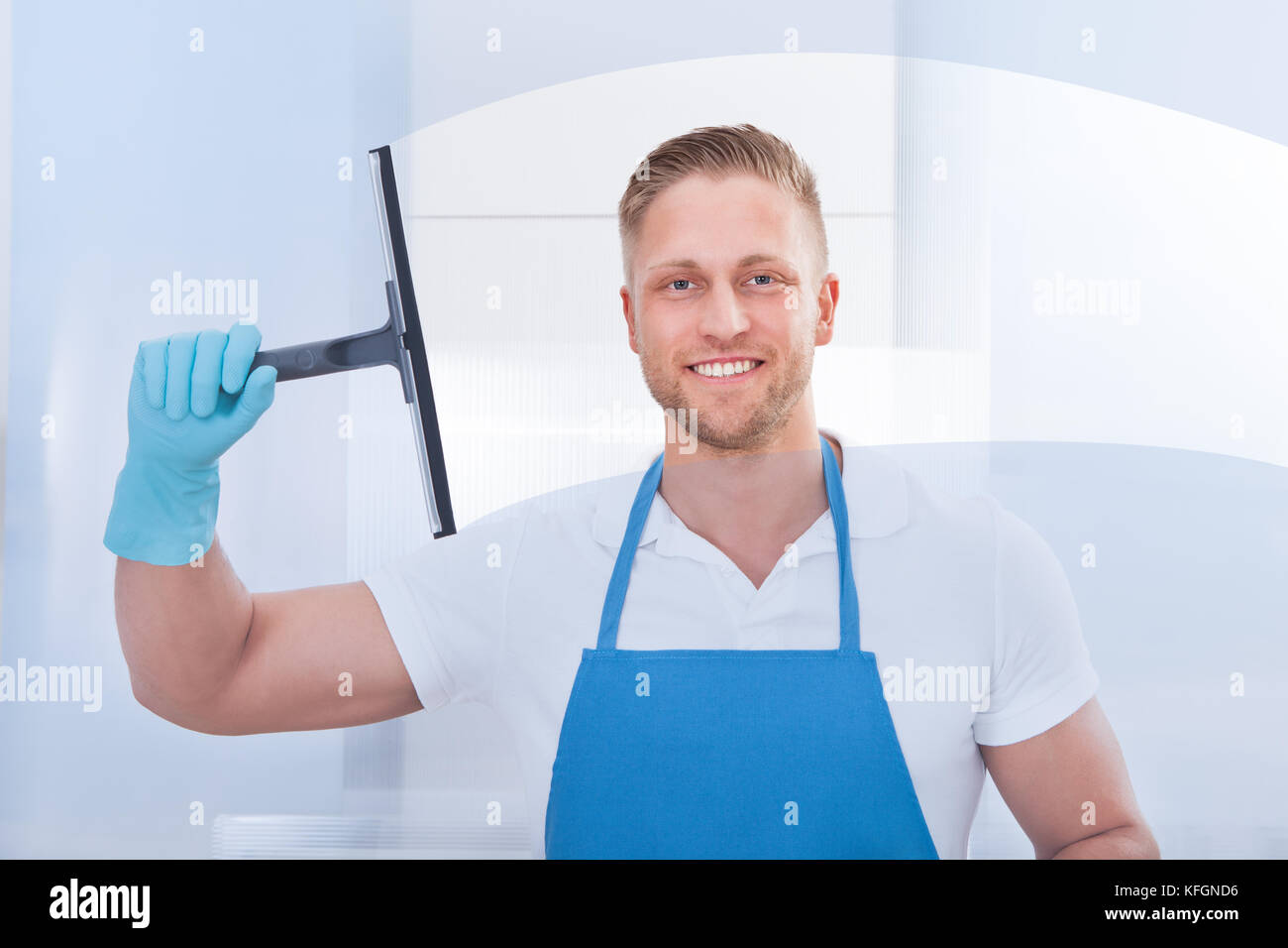 Männliche Hausmeister mit einem Rakel ein Fenster in einem Büro das Tragen einer Schürze und Handschuhe zu reinigen, da er arbeitet Stockfoto