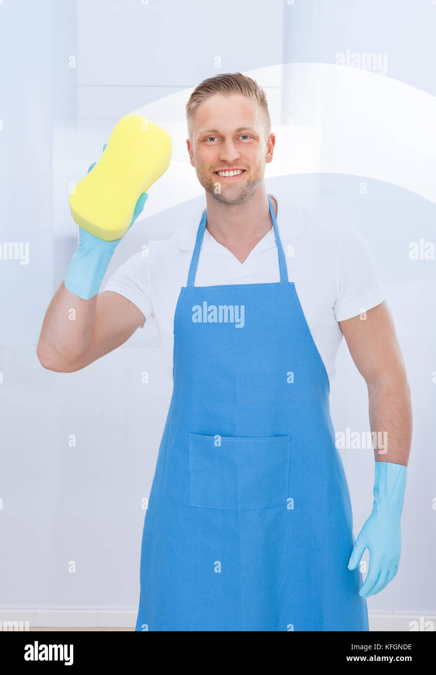 Männliche Hausmeister mit einem Schwamm ein Fenster in einem Büro das Tragen einer Schürze und Handschuhe zu reinigen, da er arbeitet Stockfoto
