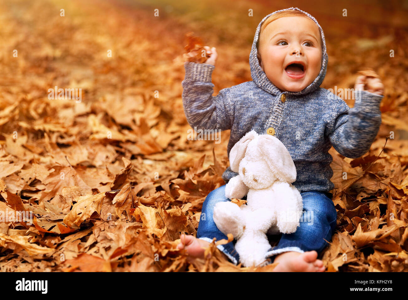Süße kleine Baby Junge sitzt auf dem Boden mit trockenem Laub im Park bedeckt, wenig fröhliches Kind spielt mit Soft Toy im Herbst Garten Stockfoto