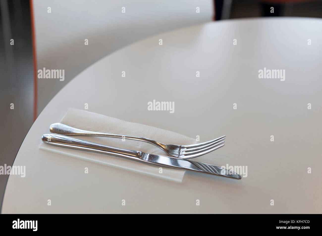 Silbermesser und Gabel auf weißer Papierserviette auf dem weißen runden Tisch mit verschwommener Stuhlrückseite im Hintergrund Stockfoto
