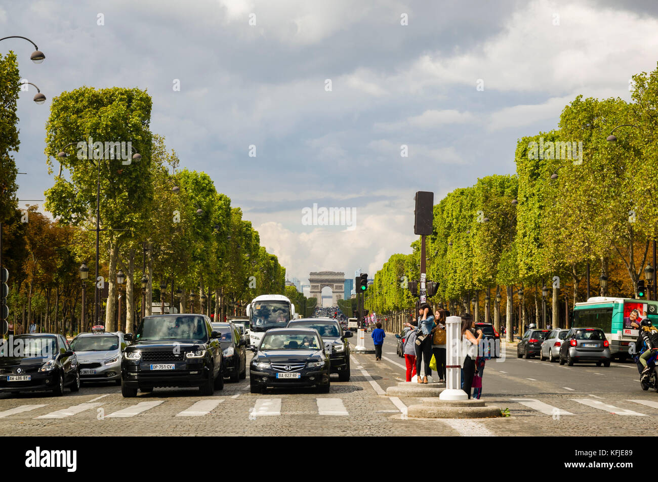 Autos gestoppt an der Ampel, auf den von Bäumen gesäumten Champs-Elysee in Paris, Frankreich, Menschen warten auf einen Zebrastreifen überqueren, der Triumphbogen im Hintergrund unter einem blauen bewölkten Himmel. Kopieren Sie Platz. Stockfoto