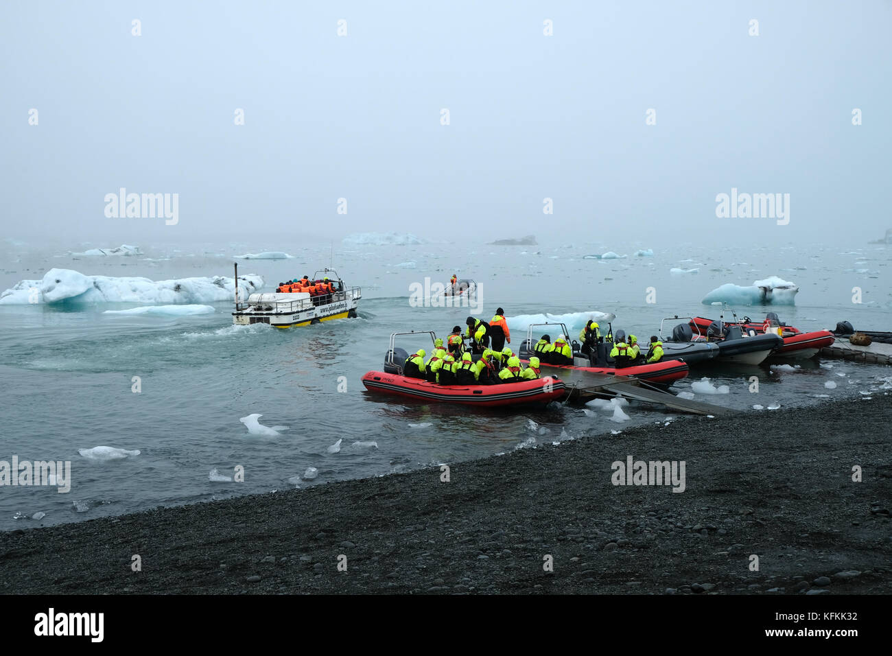 Touristen in Tierkreisbooten, die das Ufer verlassen, auf der Jokulsarlon Gletscherlagune, die Eisberge in leuchtend gelben Lebenswesten, Südisland, betrachtet Stockfoto