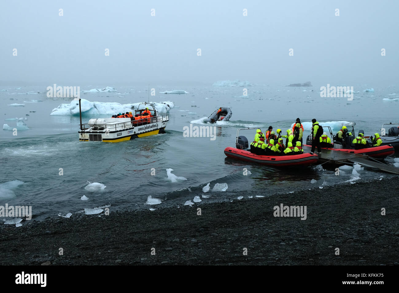 Touristen in Tierkreisbooten, die das Ufer verlassen, auf der Jokulsarlon Gletscherlagune, die Eisberge in leuchtend gelben Lebenswesten, Südisland, betrachtet Stockfoto