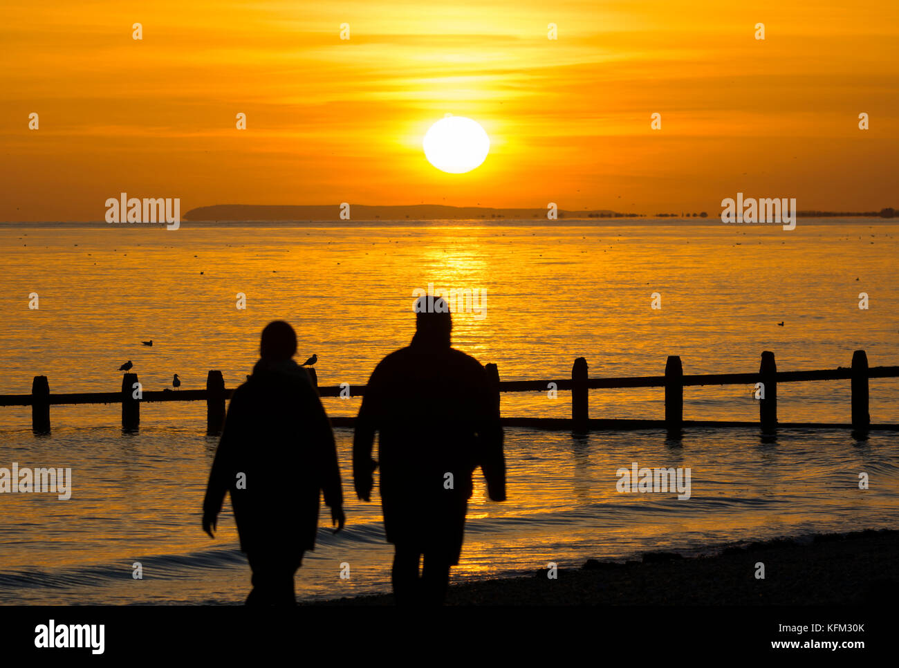 Ein Paar, das im Herbst in Großbritannien am Strand spazieren geht, während die Sonne über dem Meer untergeht. Stockfoto