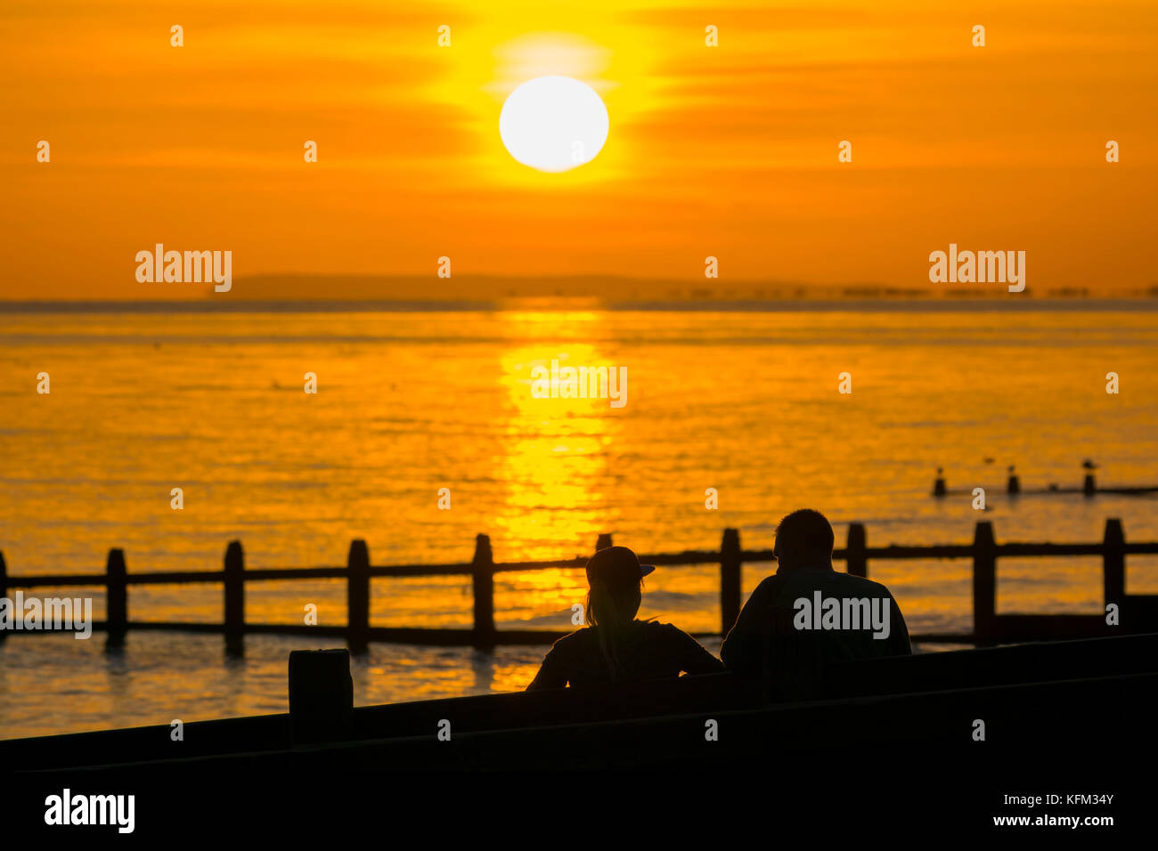 Ein Paar sitzt am Strand und beobachtet den Sonnenuntergang über dem Meer im Herbst in Großbritannien. Stockfoto