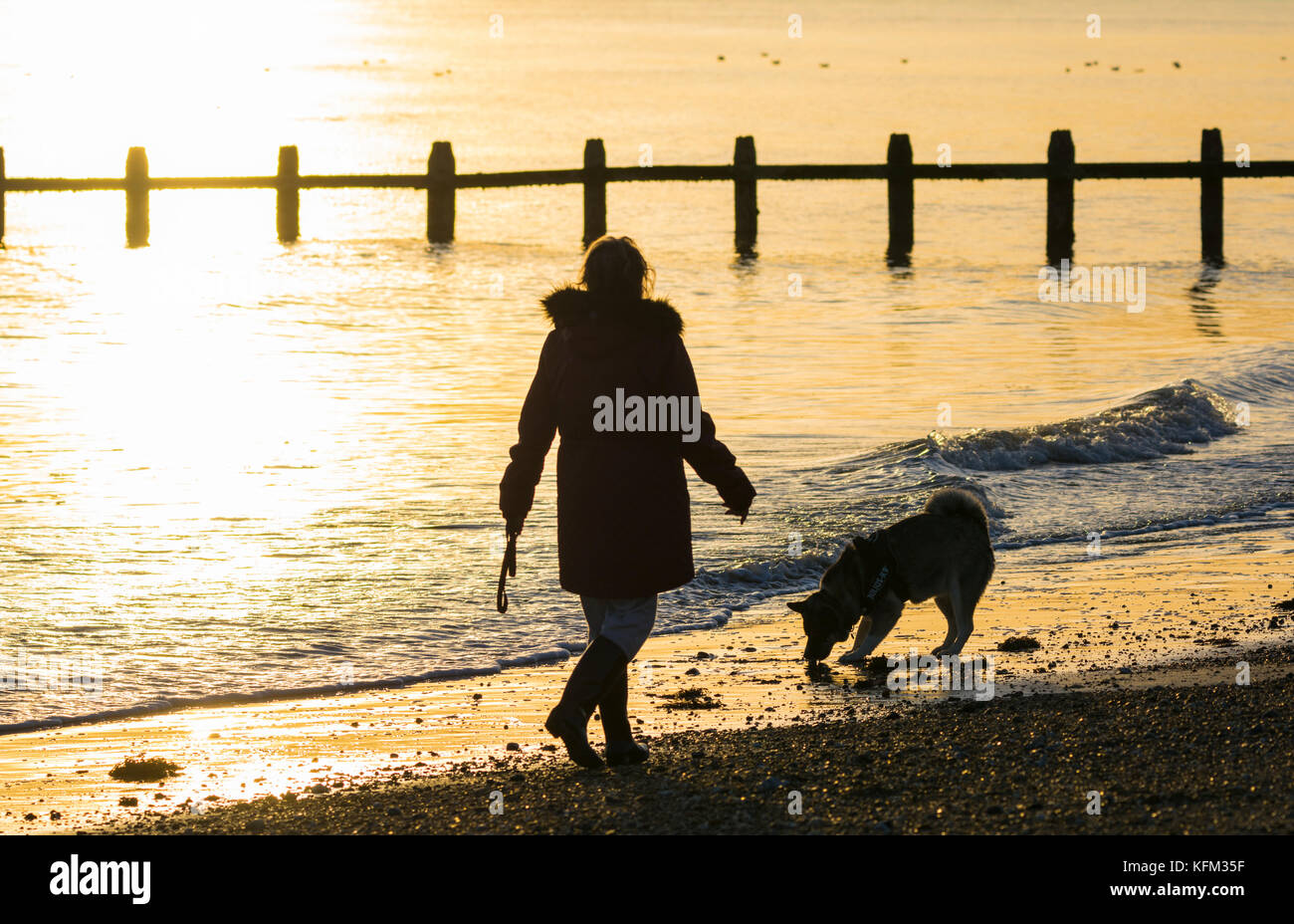 Frau, die im Herbst in Großbritannien mit einem Hund am Strand spazieren geht, während die Sonne untergeht. Stockfoto