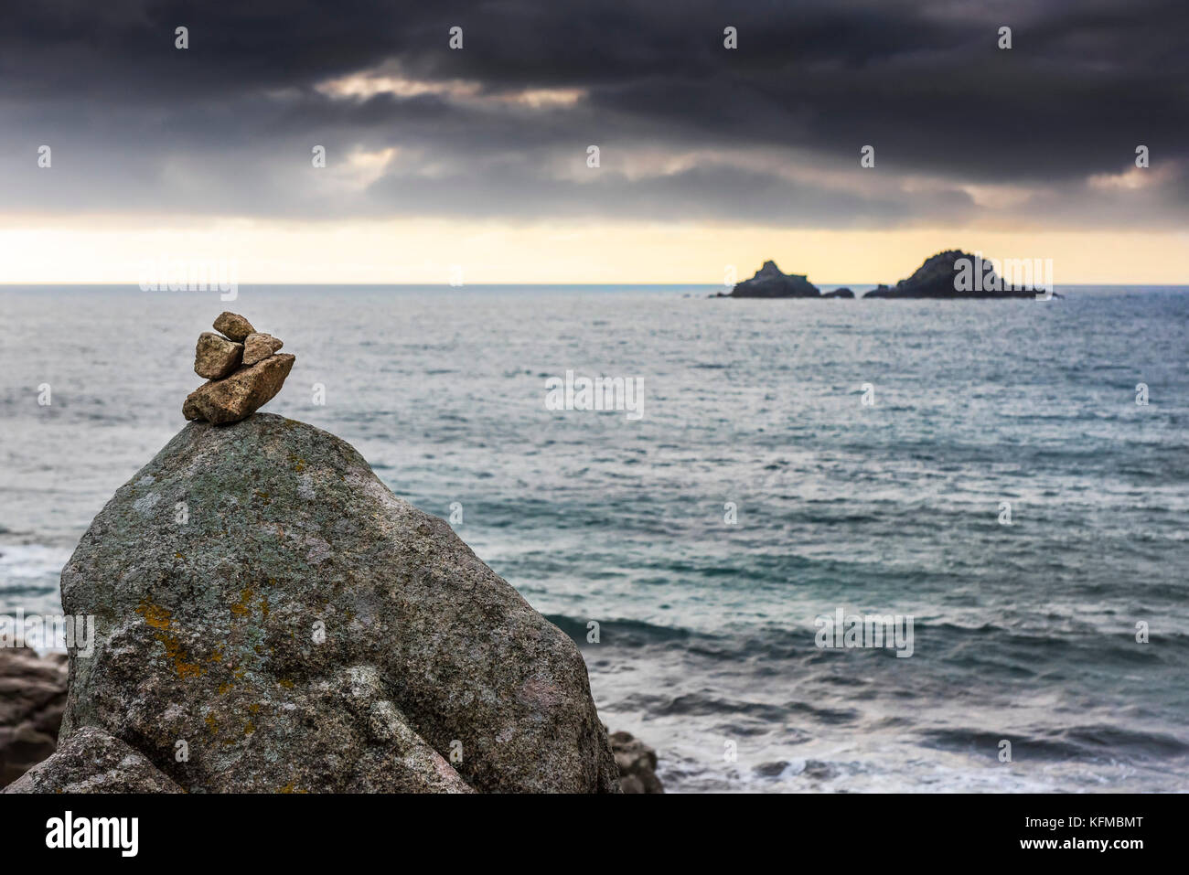 Porth Nanven Cornwall - ein kleiner Gesteinsstapel, der auf einem größeren Gestein bei Porth Nanven mit den Brisons im Hintergrund balanciert ist. Stockfoto