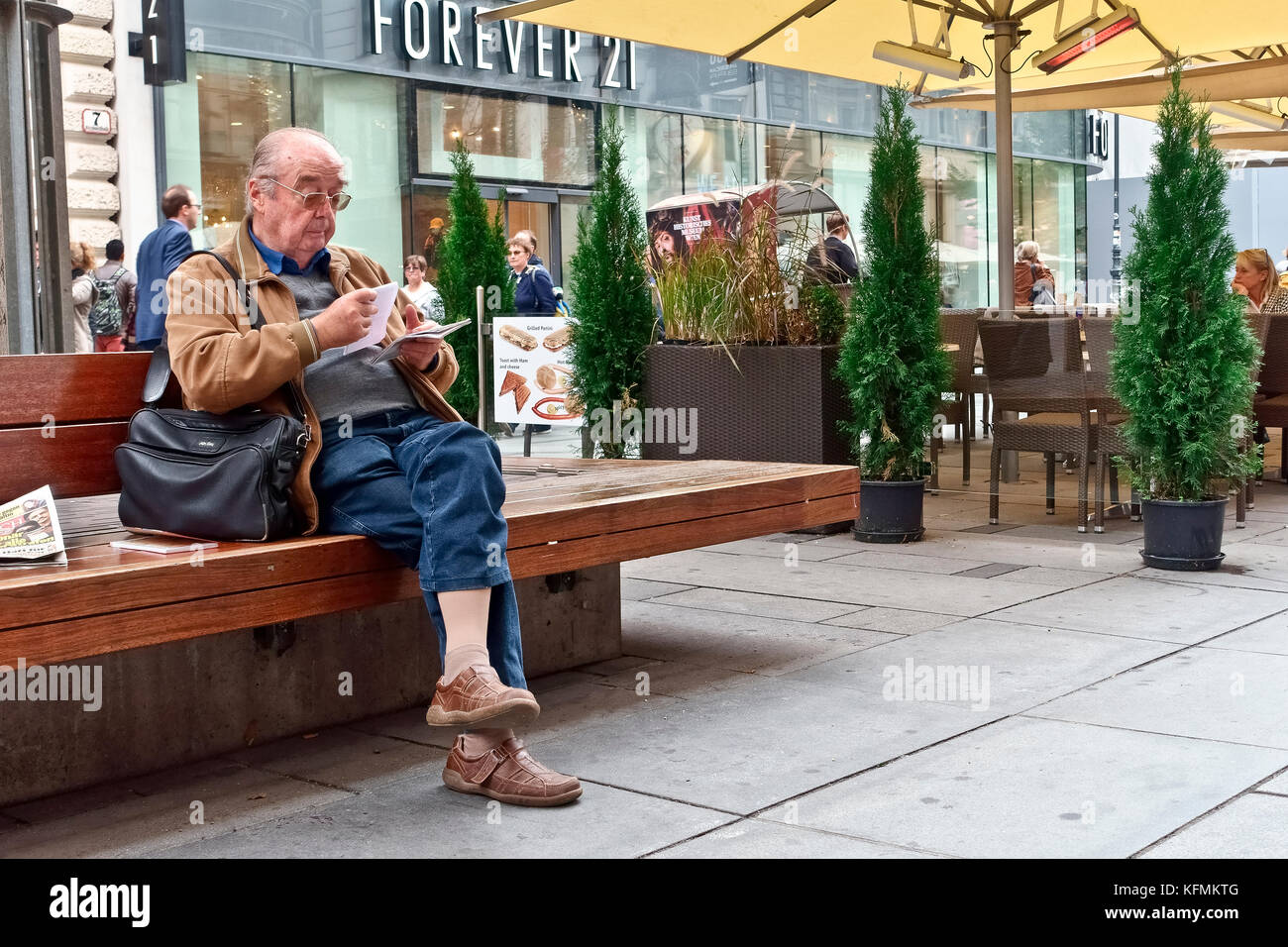 Ältere, Senior kaukasischen Mann mit Brille, alter Rentner mit gekreuzten Beinen auf einer Bank sitzen, lesen Papiere, in der Innenstadt von Wien, Österreich, Europa Stockfoto