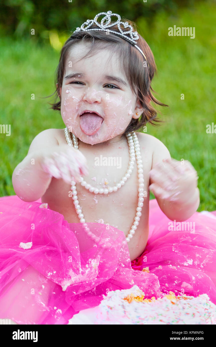 Smiling Happy Baby Kleinkind Mädchen erster Geburtstag Jahrestag Partei. Lachen und Kleben mit herausgestreckter Zunge, Gesicht schmutzig von rosa Kuchen. Princess Tiara Kostüm Stockfoto