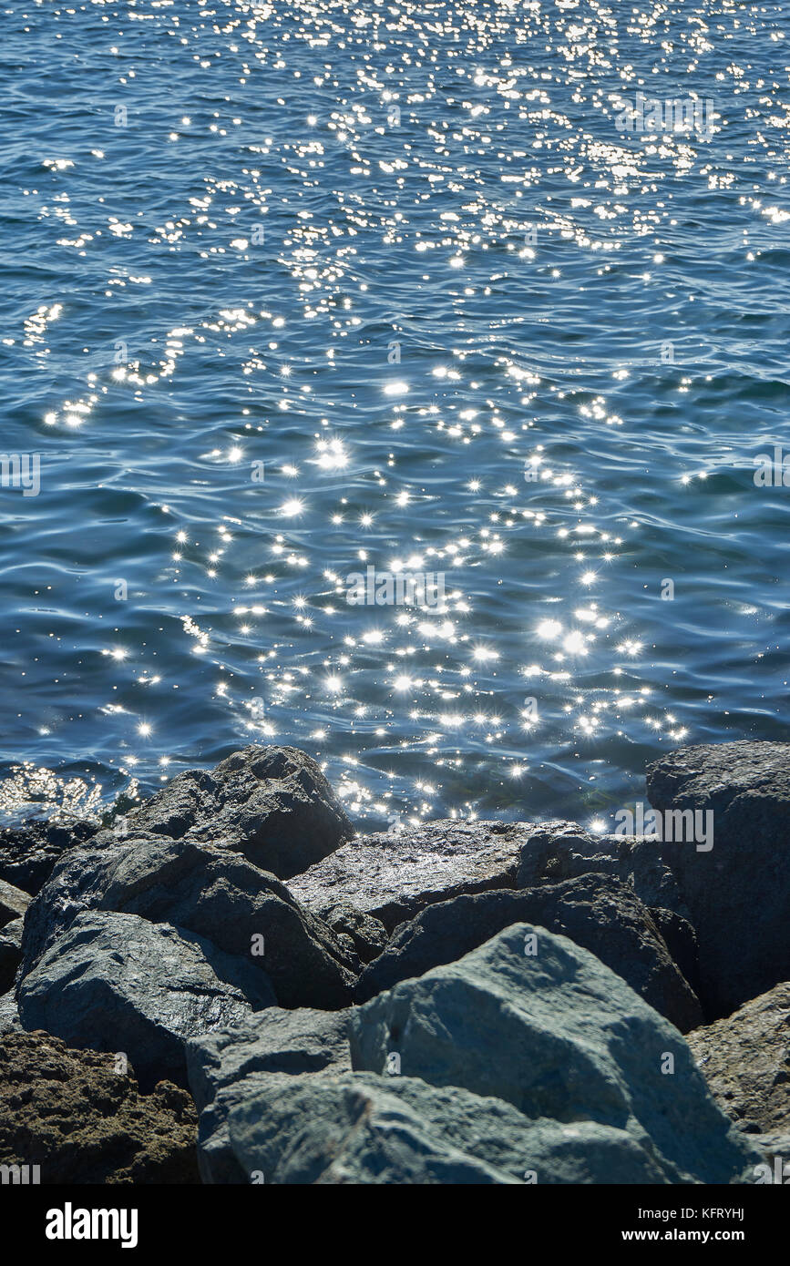 Steine und Felsen im Vordergrund der glitzernden Sonne auf dem Wasser Stockfoto