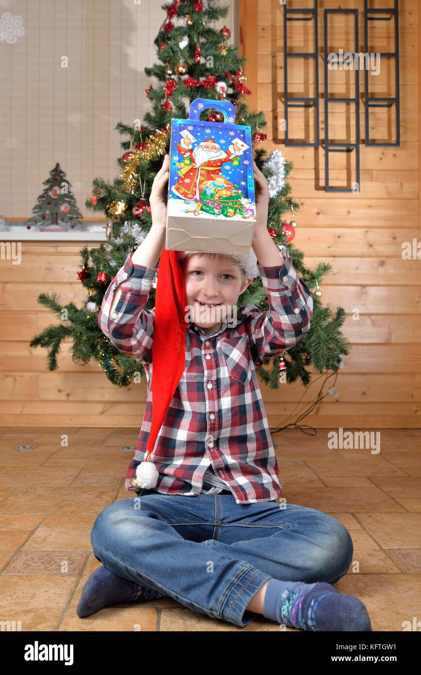 Der Junge sitzt auf dem Boden und hält in seiner ausgestreckten Händen ein Geschenk unter dem Weihnachtsbaum. Stockfoto