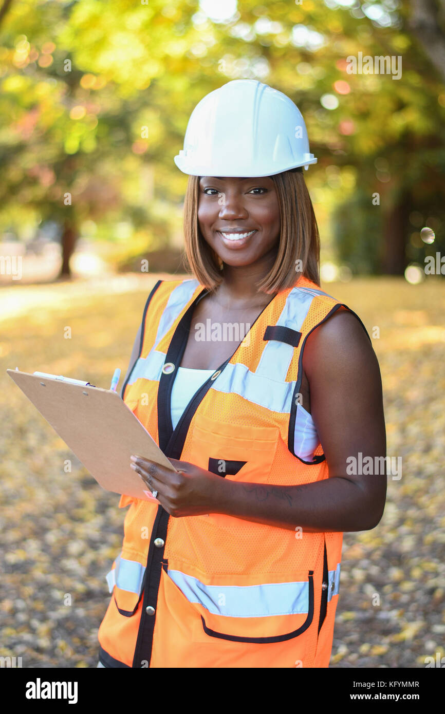 Ein schwarzes Weibchen Bauarbeiter Tragen eines weißen Hut und orange Weste außerhalb einer Stadt park arbeiten Stockfoto