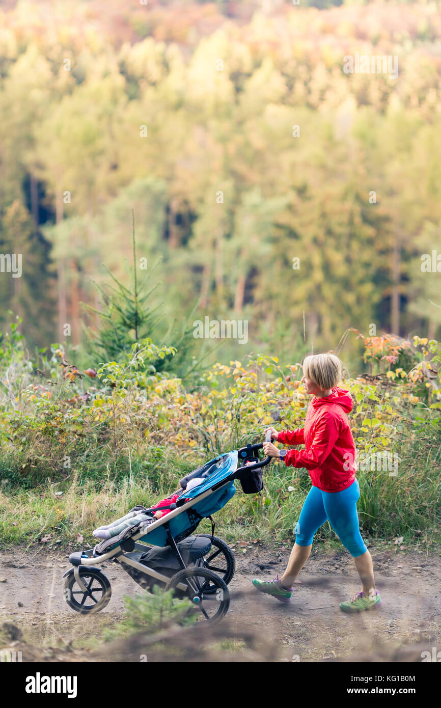 Wandern Mutter mit Kind im Kinderwagen bei Mutterschaft im Herbst Sonnenuntergang und Bergwelt. Joggen oder Walken Frau mit Kinderwagen in den Wald. Stockfoto