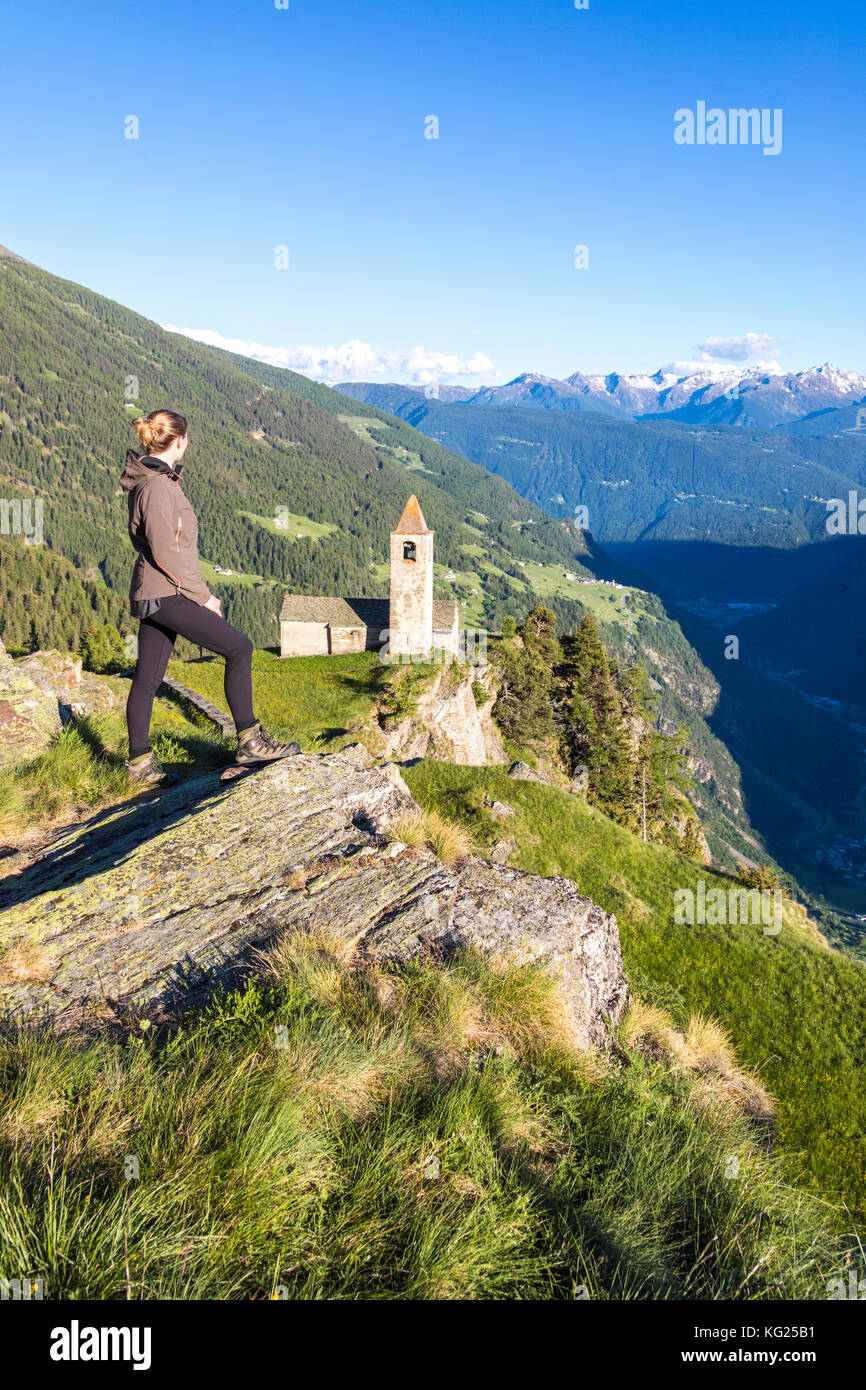 Die Frau schaut auf die alte Kirche auf den Bergen, die San Romerio Alp, Brusio, den Kanton Graubünden, das Poschiavo-Tal, Schweiz, Europa Stockfoto