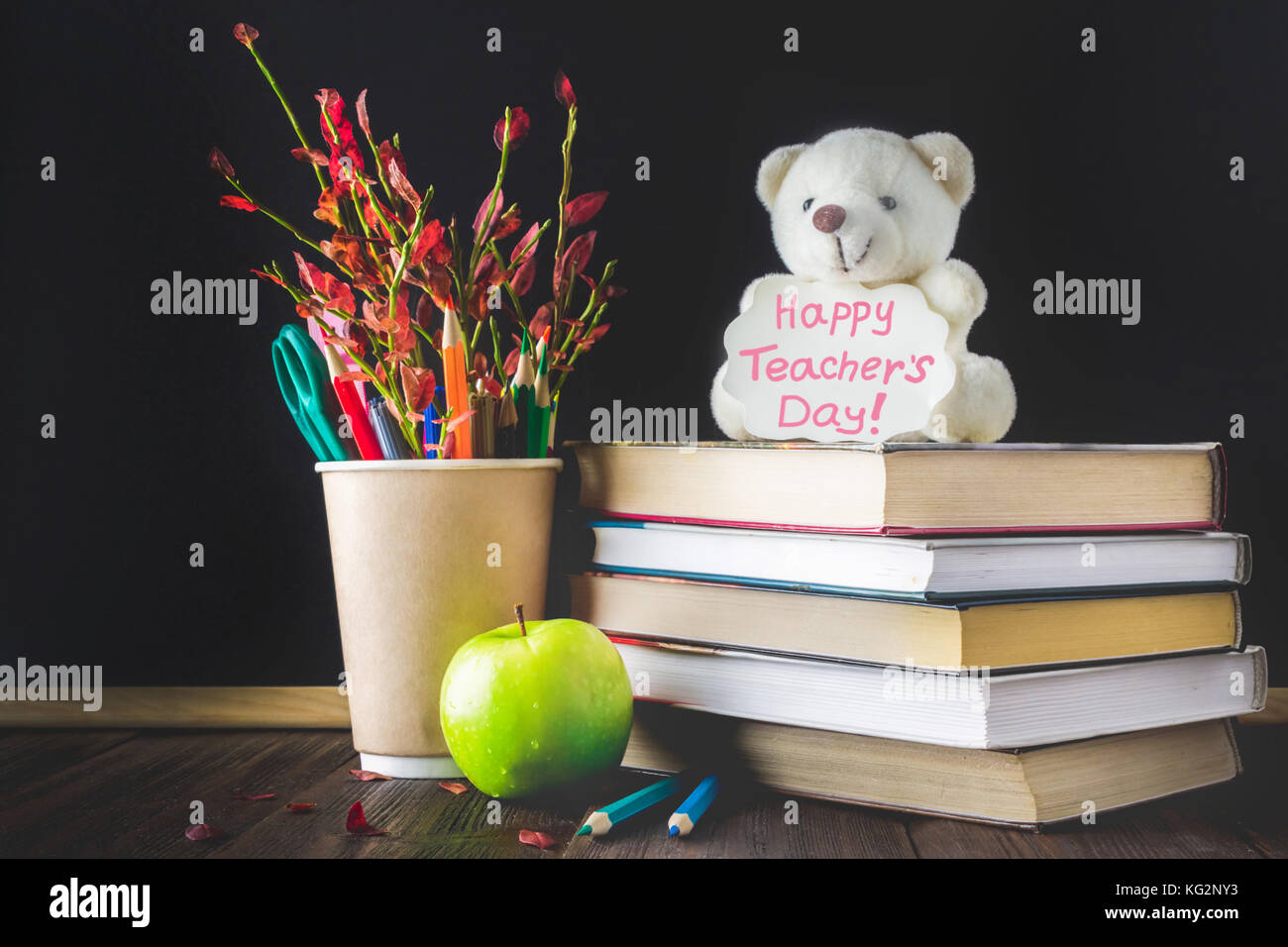 Konzept der Lehrer Tag. Objekte auf einer Schiefertafel Hintergrund. Bücher, grüner Apfel, Bär mit einem Schild: happy Lehrer Tag, Bleistifte und Kugelschreiber in einem Glas, Stockfoto