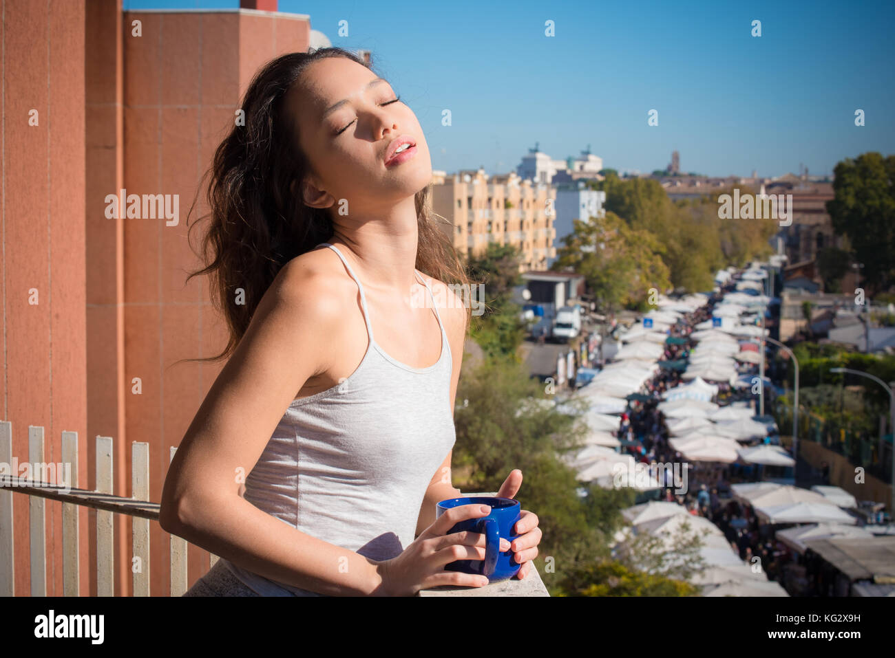 Jungen asiatischen Teenager Frau auf dem Balkon hoch geschlossenen Augen Sonnenlicht Blau Becher in die Hände städtischer Hintergrund Stadt Lebensstil Stockfoto