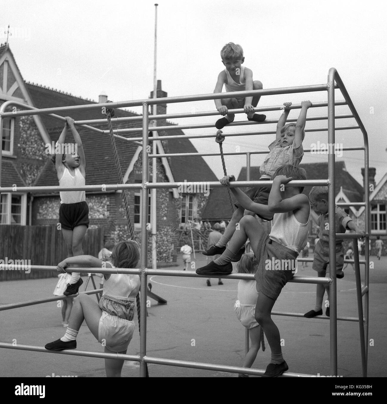 In den 1950er Jahren spielen historische Jungen und Mädchen aus dem Kindergarten auf einem Metallkletterrahmen draußen in ihrer Dorfschule auf dem Spielplatz und machen eine PE-Klasse, einige davon in ihrem Gym-Kit, England, Großbritannien. Stockfoto