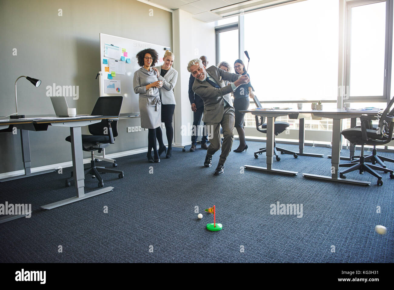 Vielfältige Gruppe von Lachen Mitarbeiter beobachten ein reifer Kollege Schwingen einer Toy Golf Club während einer Übung in einem modernen Büro Stockfoto