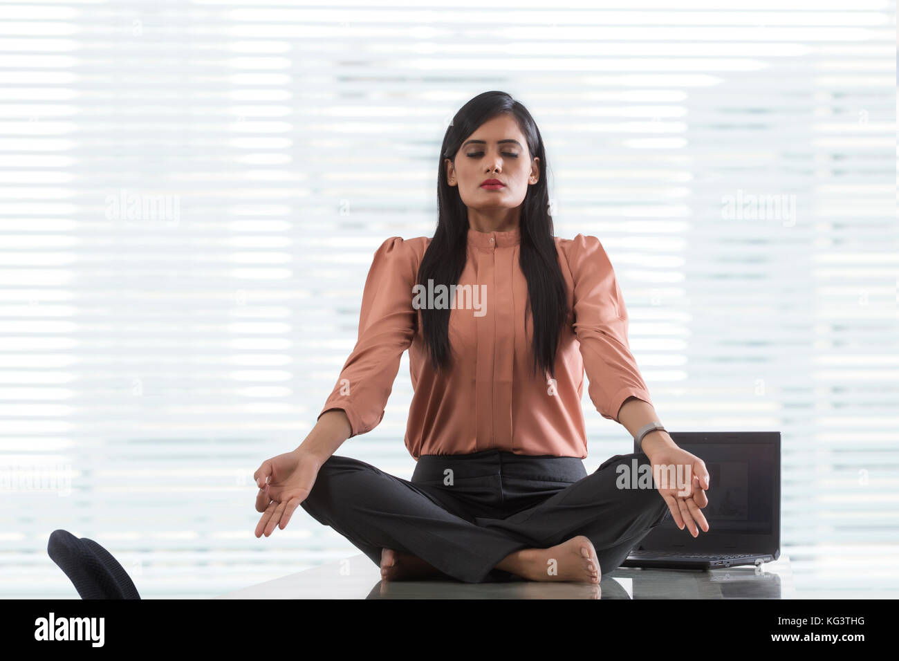 Junge Geschäftsfrau mit überkreuzten Beinen Yoga auf dem Schreibtisch im Büro Stockfoto
