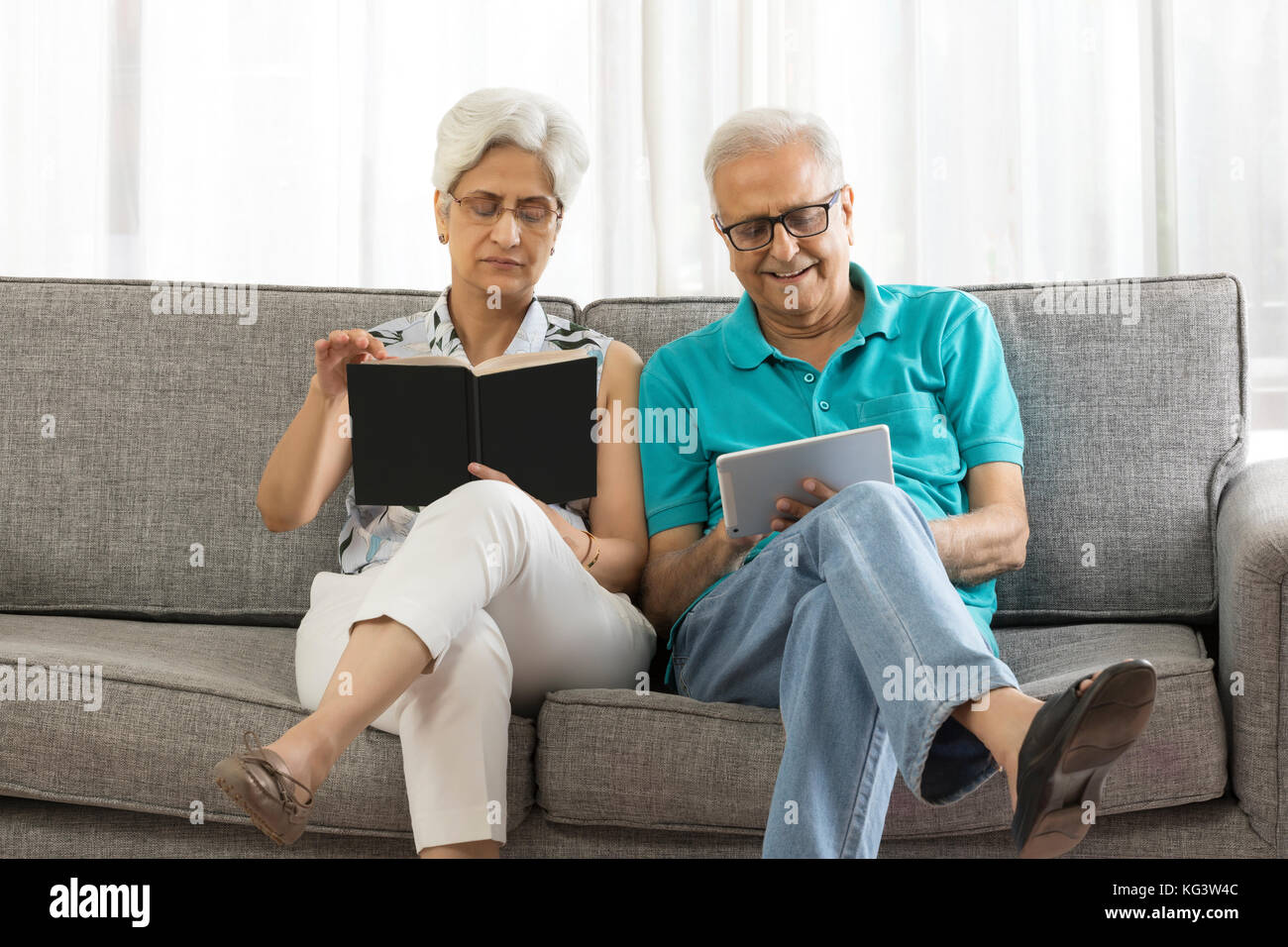 Ältere Frau mit Buch und älterer Mann mit digitalen tablet Sitzen auf einem Sofa Stockfoto