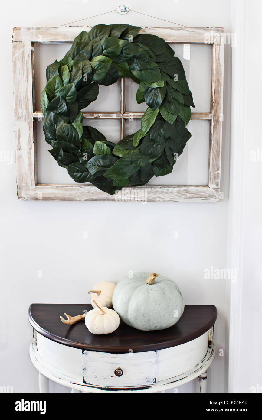 Altes Bauernhaus Fenster mit einem hausgemachten magnolia Blatt Kranz an einer Innenwand über rustikale half moon Tabelle mit erbstück Kürbisse aufgehängt eingerichtet. Stockfoto