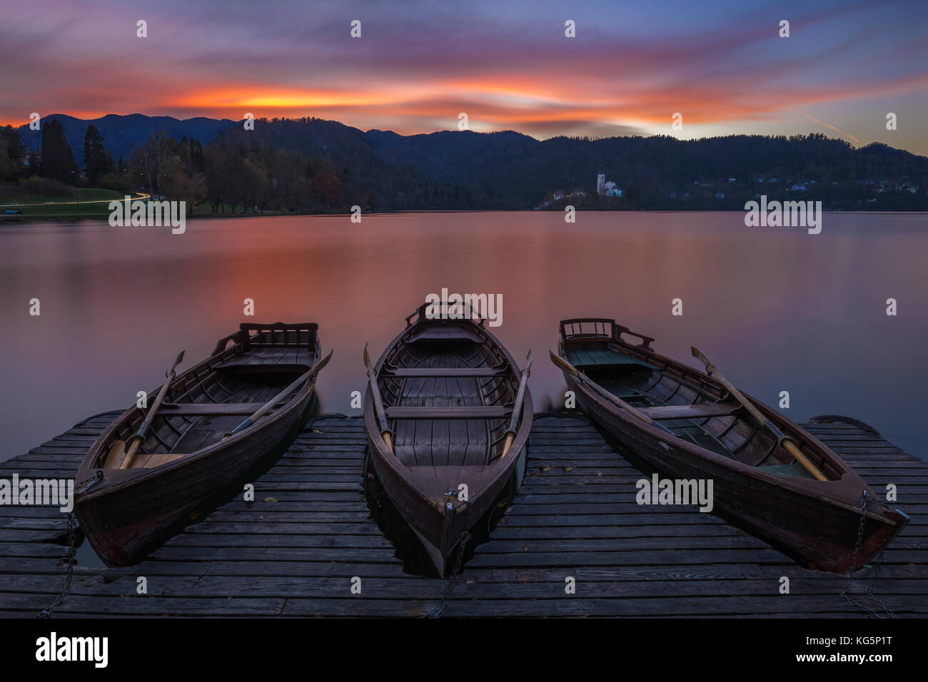Bled, Slowenien - Traditionelle Boote am Bleder See mit wunderschönem, dramatischem Sonnenuntergang und der Maria-Pilgerkirche und den Bergen am Rücken Stockfoto