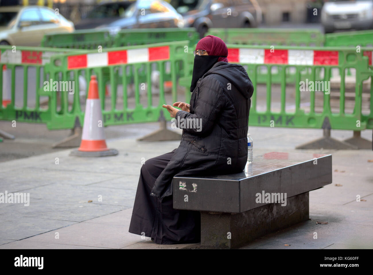 Asiatische Frauen Mädchen Flüchtling Hijab Schal auf der Straße in Großbritannien muslimische moslem mit Smartphone sitzend auf Bank Straße mit Warnung Verkehrsbarrieren Gefahr Stockfoto