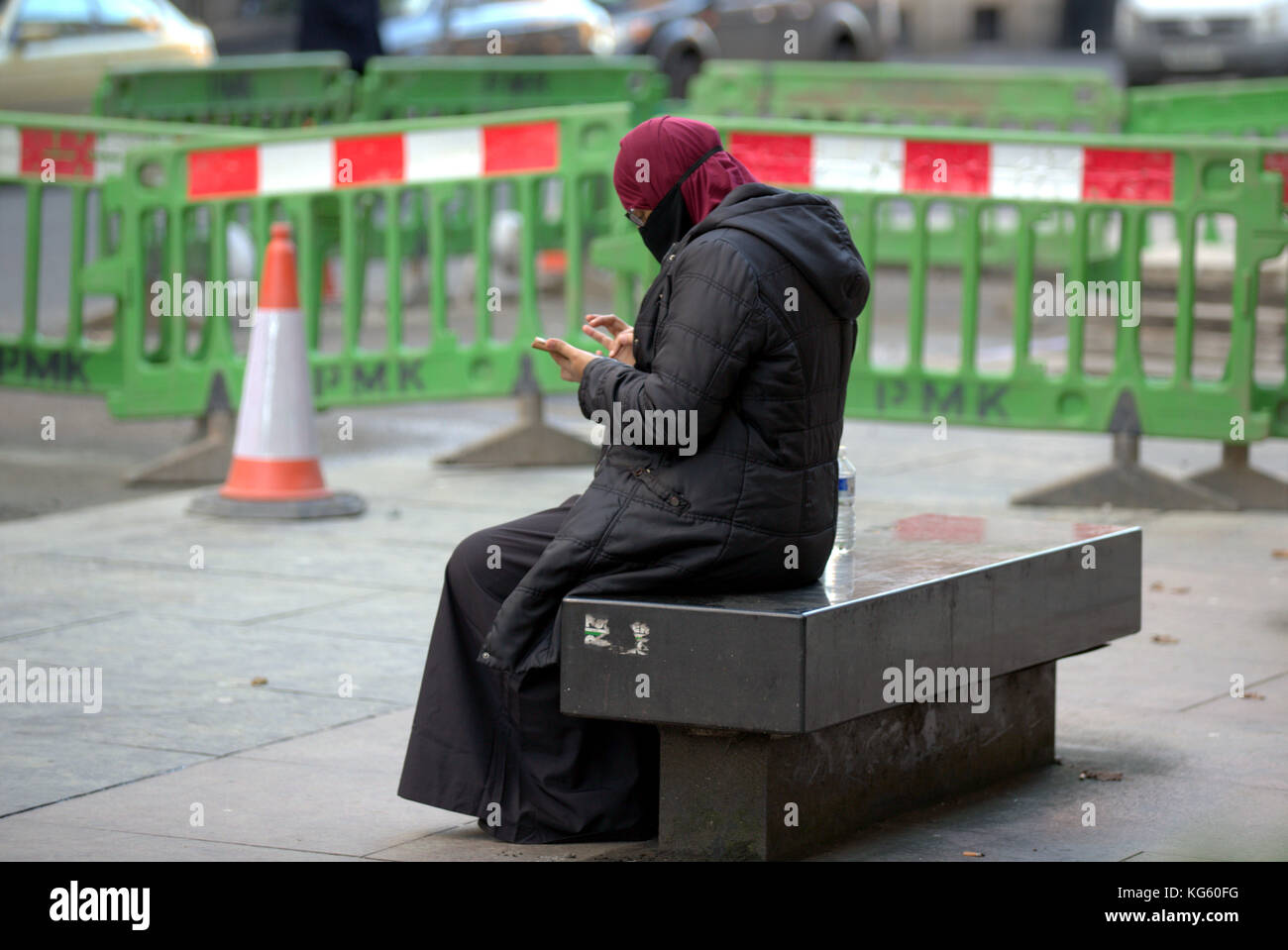 Asiatische Frauen Mädchen Flüchtling hijab Schal auf der Straße in der britischen muslimischen moslemischen mit Smartphone sitzt auf der Bank Street mit Warnung Verkehr Barrieren Gefahr Stockfoto