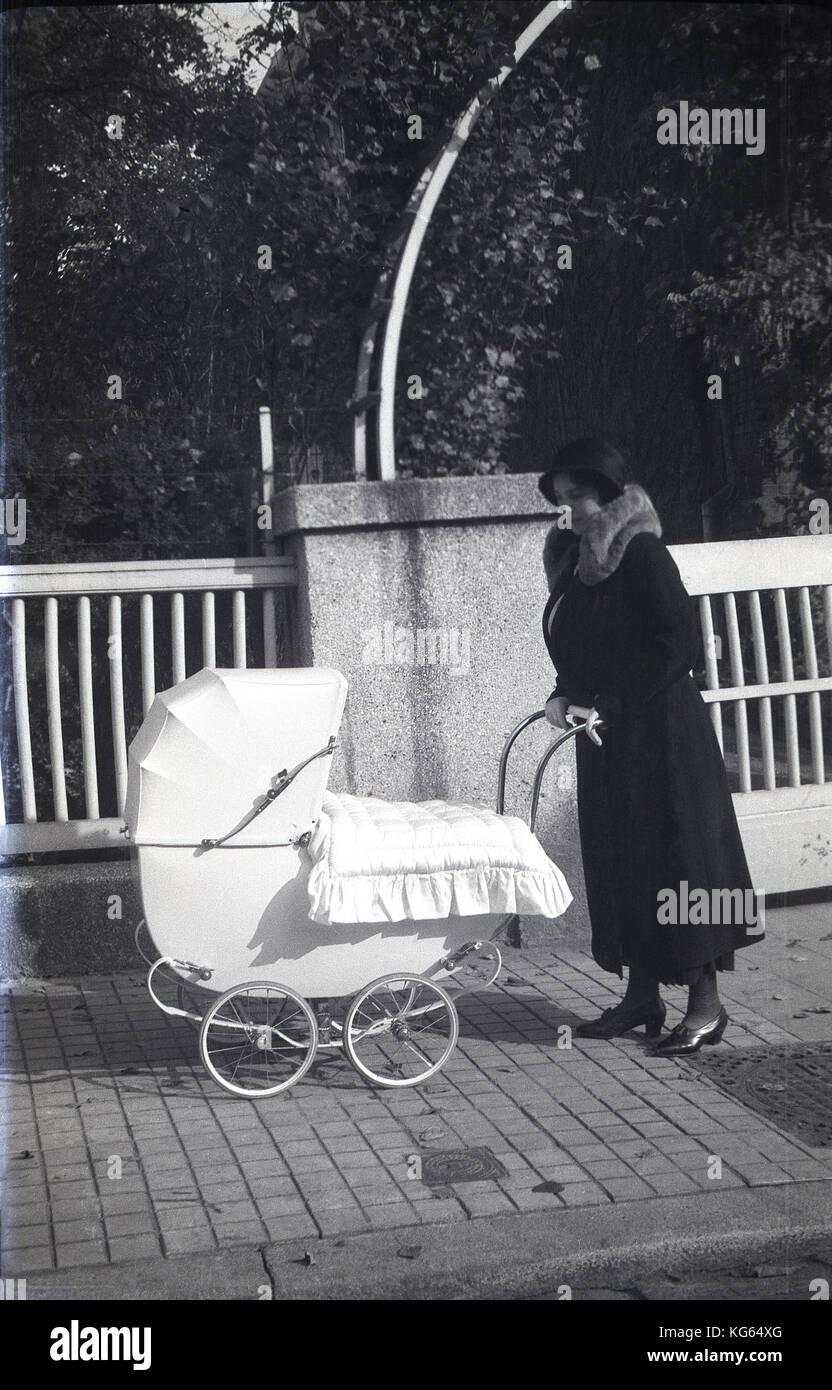 @ 1920 s, einem modischen jungen Mutter des Tages trägt einen pelzbesatz Mantel und Hut mit ihrer stilvollen Federbelastete vierrädrigen Kinderwagen außerhalb auf einem Gehsteig mit einer Decke abgedeckt werden. Stockfoto