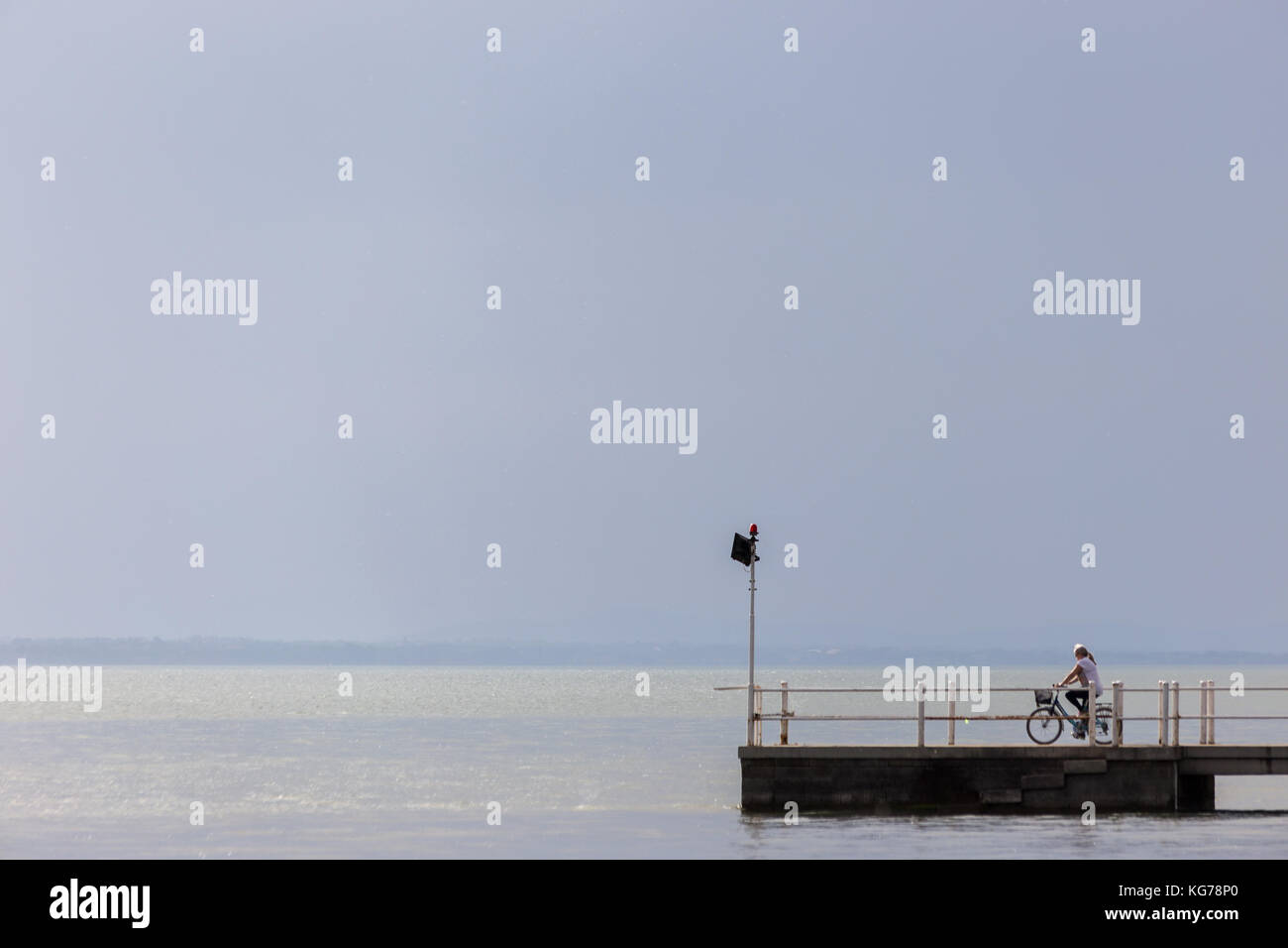 Eine Frau auf einem Fahrrad über einen Steg am See, unter einem großen leeren Himmel Stockfoto