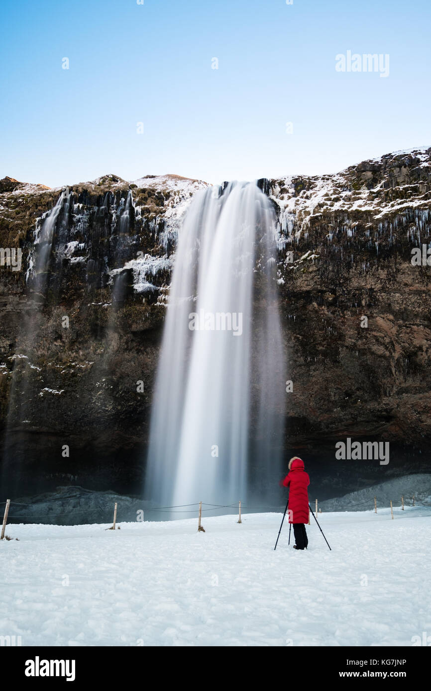 Wasserfall im Winter, Fotograf in roter Jacke, die alleine stehen auf dem Schnee am Wasserfall Seljalandsfoss in Island Stockfoto