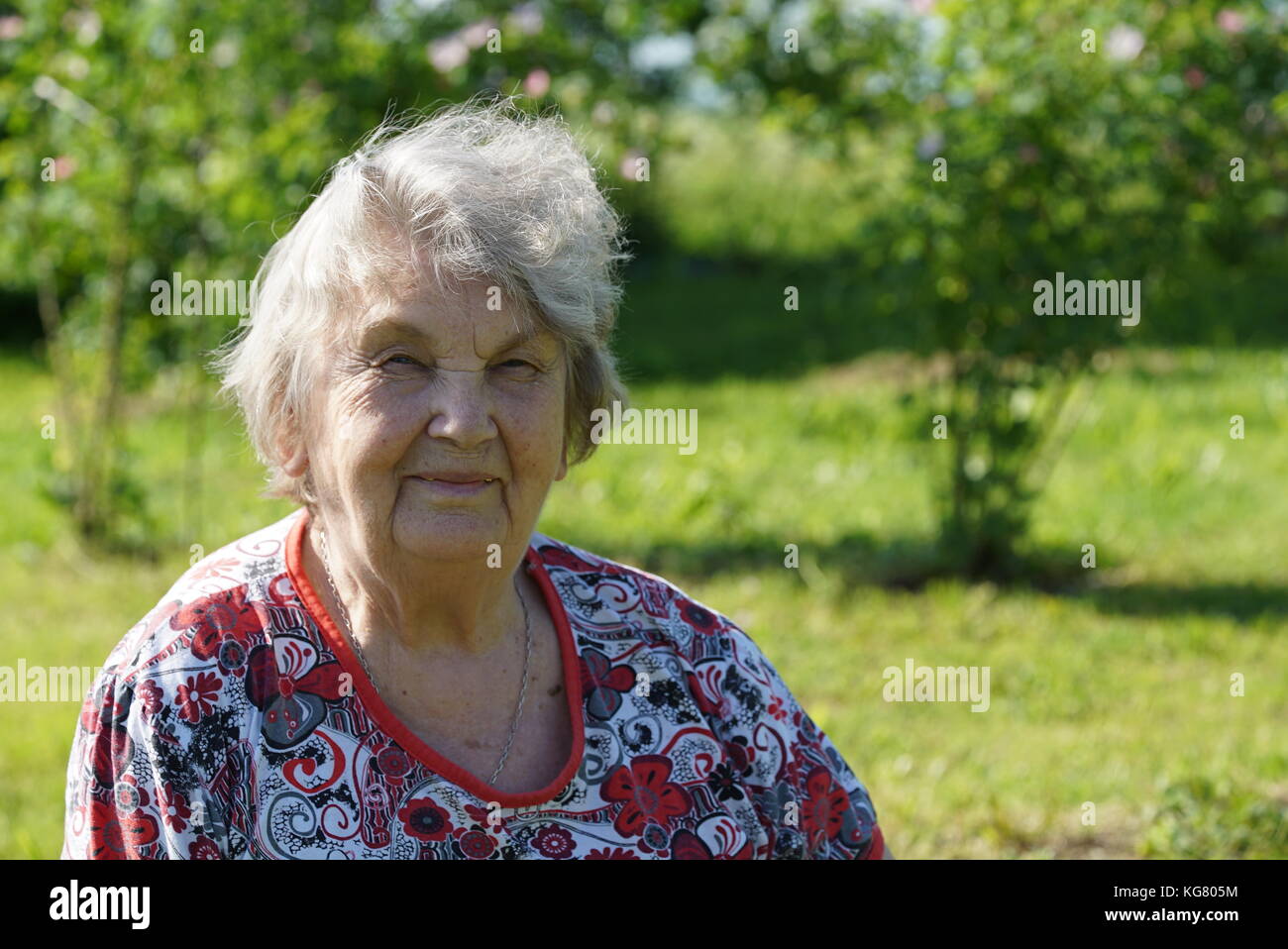 Porträt der alten Frau gekleidet in t-shirt in der gargen, die im Hintergrund von wachsenden Bäumen im sonnigen Sommertag. Frau aufgeladen ist Energie der Natur Stockfoto