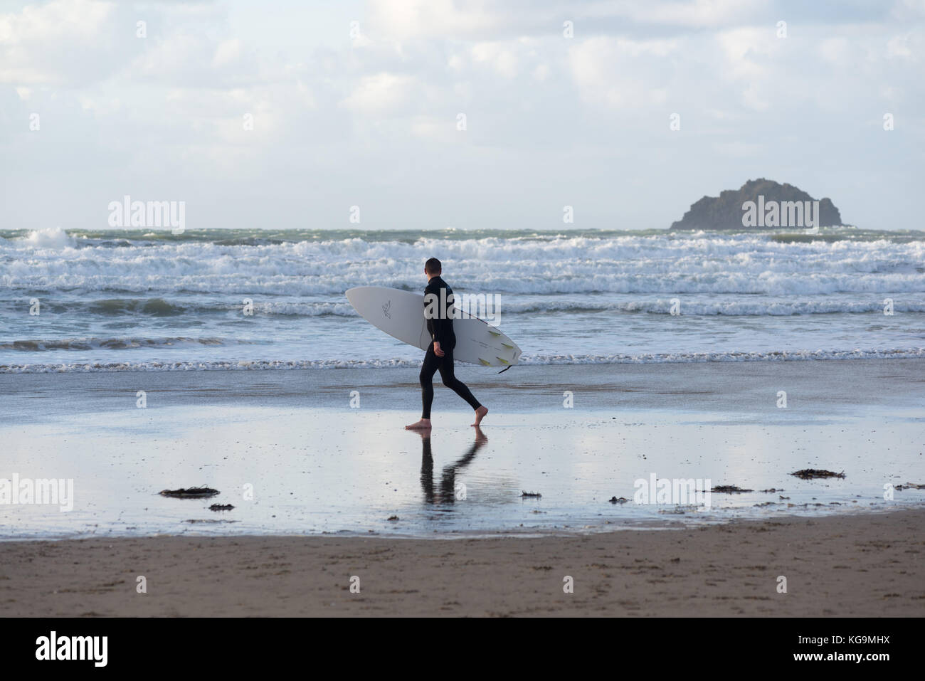 Starke Winde und kalten Wetter an der Küste von North Cornwall in hellen Herbst Sonnenschein an Polzeath, Cornwall, UK. Ein Surfer trotzt der Kälte zu Fuß am Strand entlang, seine Surf Board. Stockfoto
