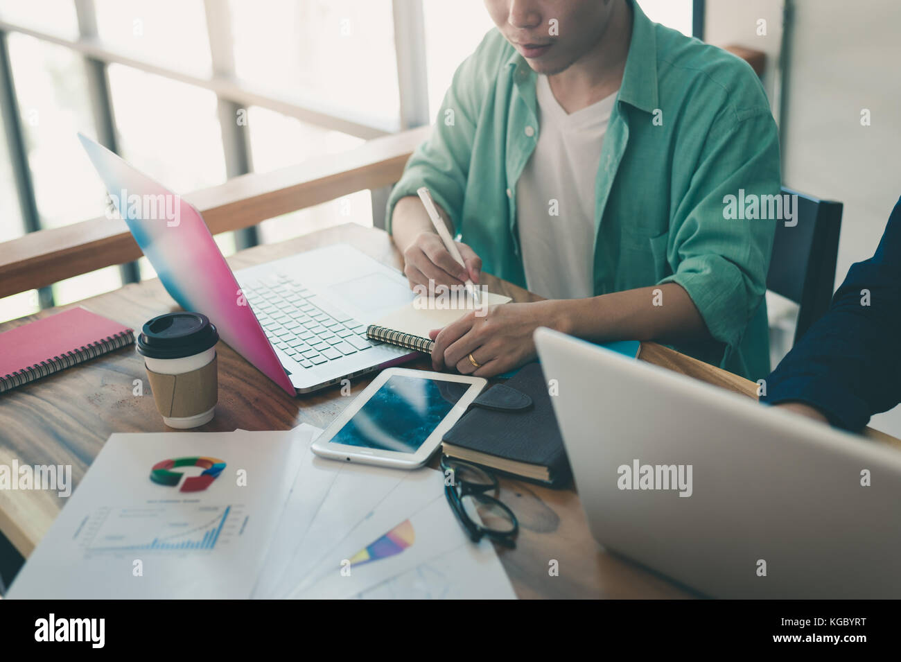 Jungen asiatischen freier Mitarbeiter schreiben auf kleine Notebook bei der Arbeit mit Kollegen im Coffee Shop. Freiberufler auf Arbeitstag Aktivität in Coworking Space Stockfoto