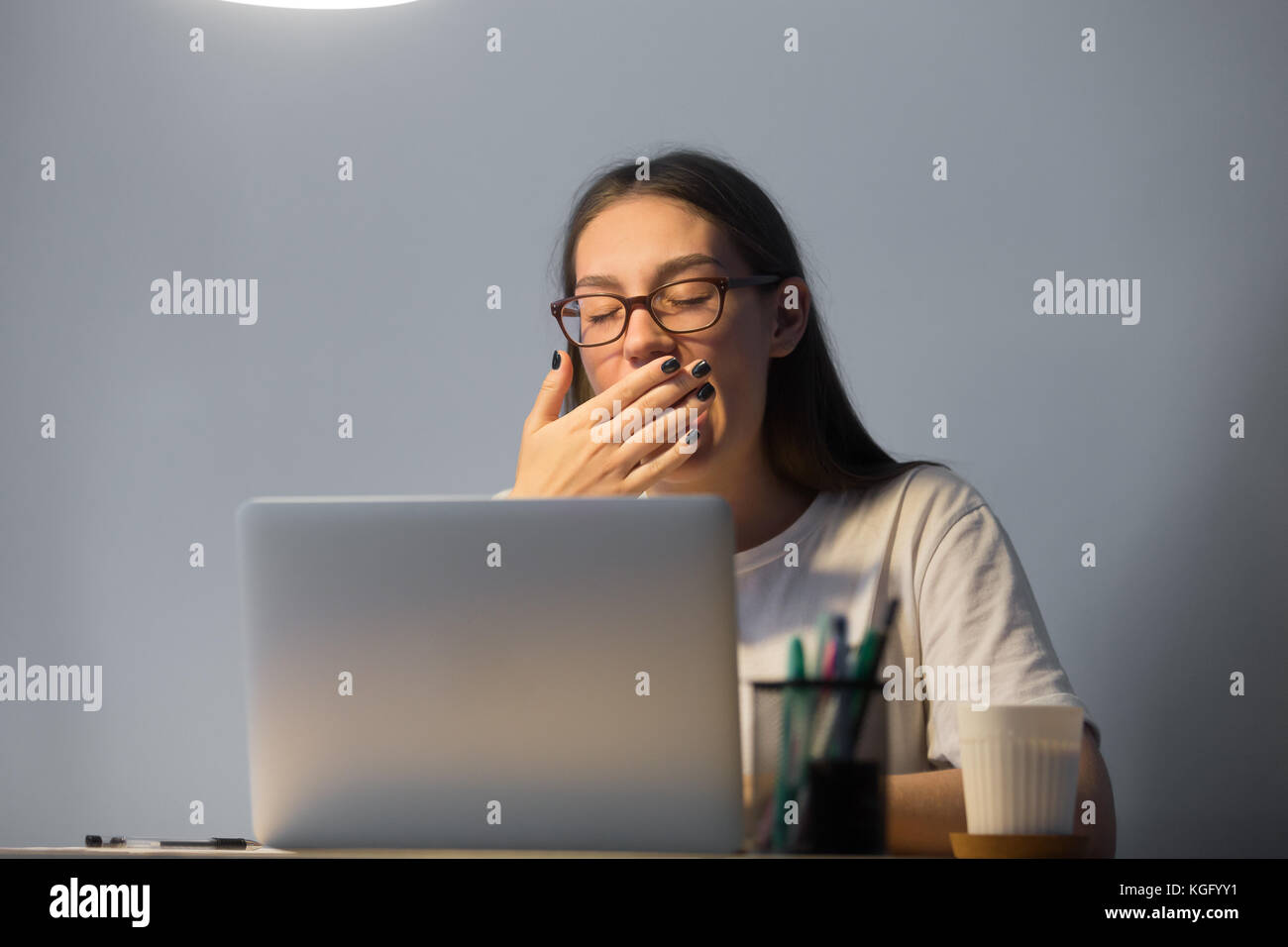 Müde junge erwachsene Frau Sekretär in Gläsern funktioniert auf dem Laptop Computer am Arbeitsplatz Schreibtisch spät in der Nacht, Gähnen, deckt den Mund mit der Hand. Stockfoto