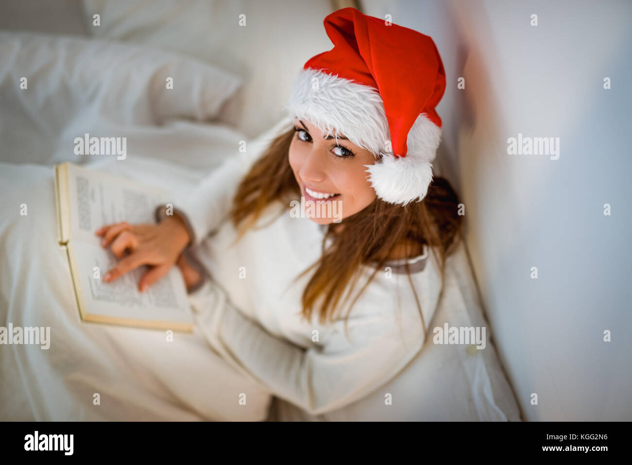 Ansicht von oben eine schöne lächelnde Mädchen mit weihnachtsmütze Entspannen in gemütlichen Bett und Lesen Buch. Bei Kamera schaut. Stockfoto