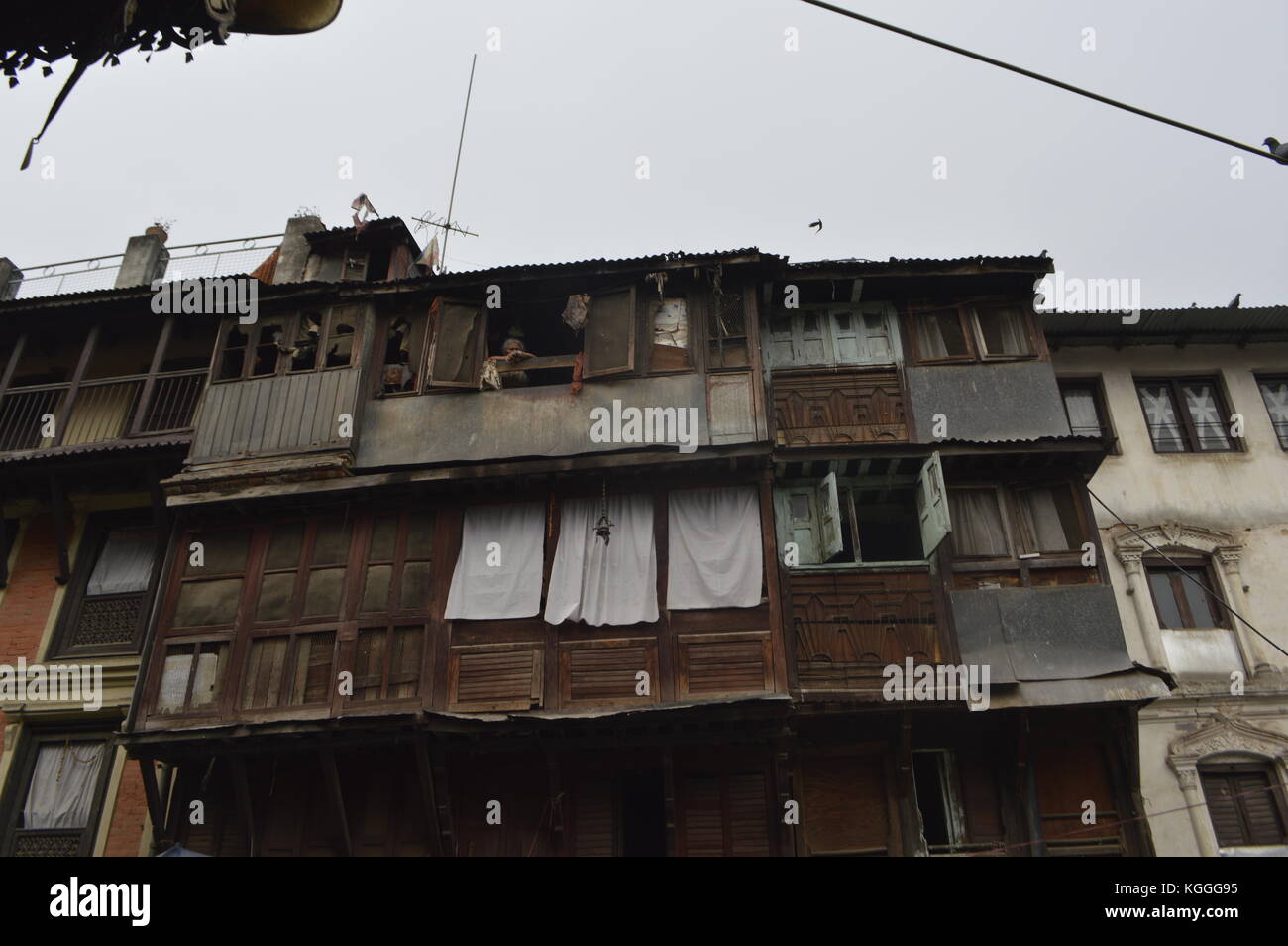 Eine alte nepalesische Dame, die in einem Holzhaus mit einer Zusammenstellung von Fenstern und Fensterläden in Kathmandu, Nepal, aus dem Fenster auf die Straße starrt. Stockfoto