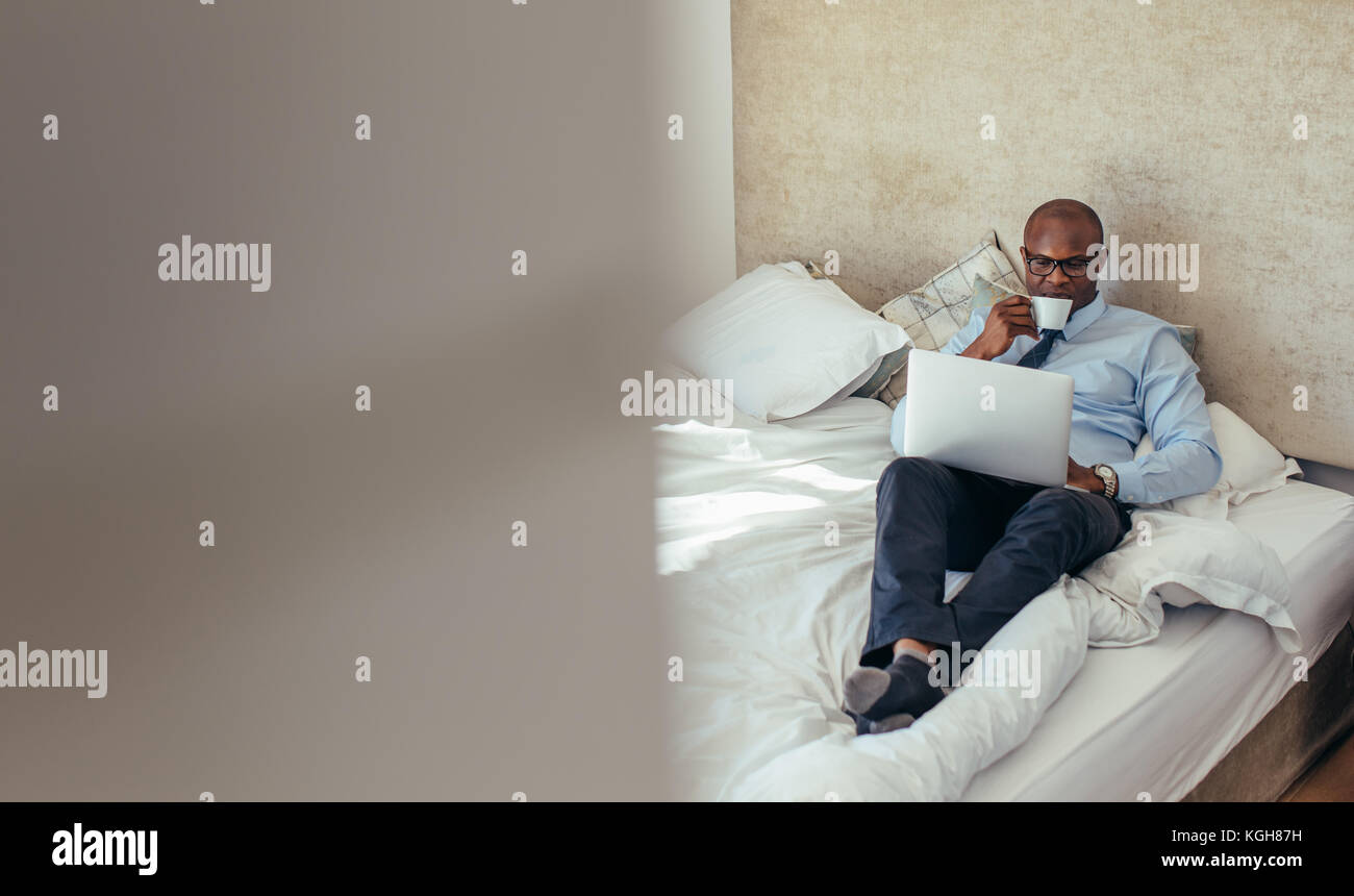 Mann in formeller Kleidung, der am Laptop arbeitet, während er im Bett liegt. Geschäftsmann, der an einem Laptop arbeitet, während er Kaffee in seinem Hotelzimmer trinkt. Stockfoto