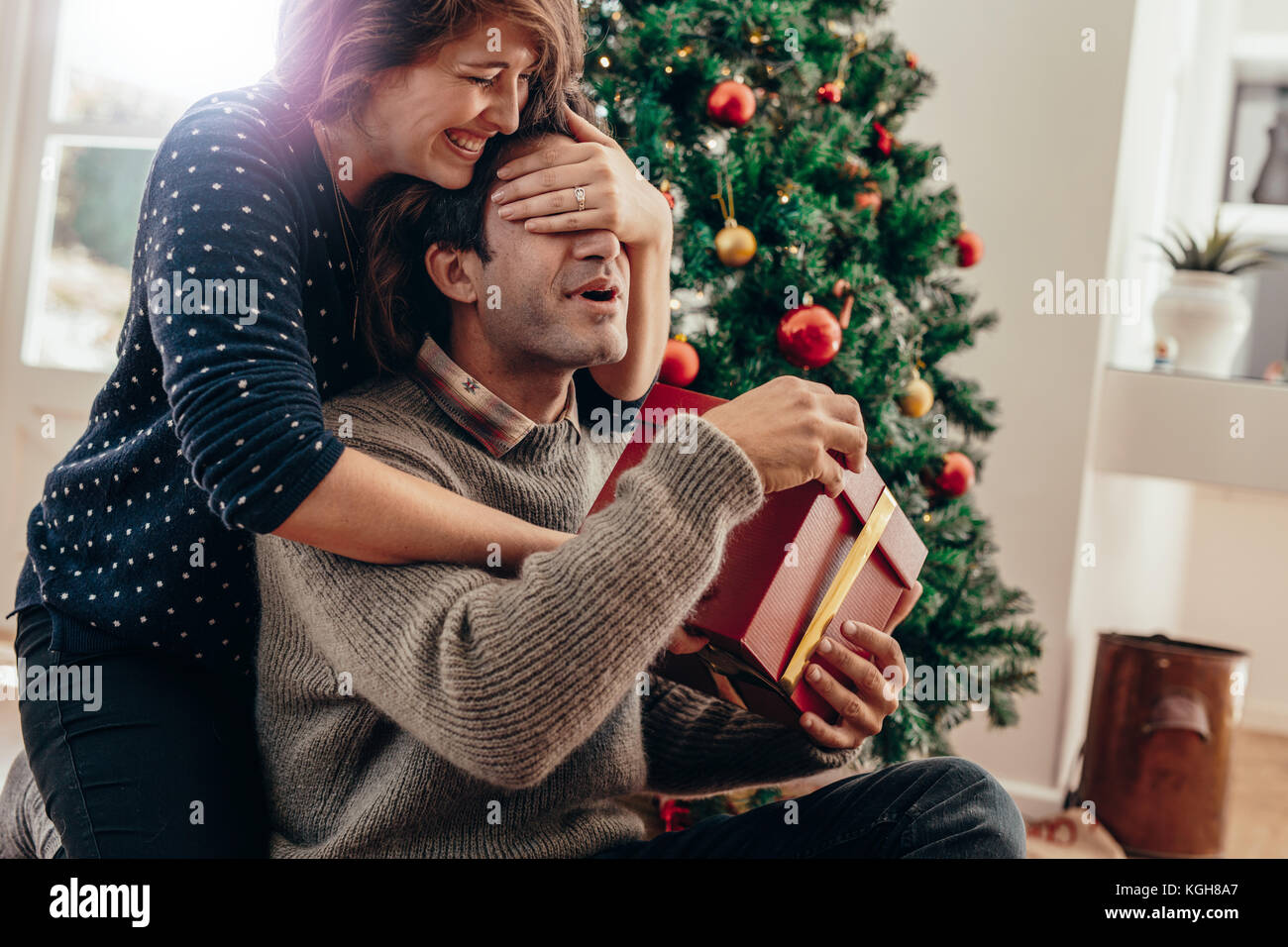 Ein paar glückliche Momente verbringen während Weihnachten zu Hause feiern. Lächelnden jungen Frau die Augen ihres Partners während über Weihnachten gif Übergabe Stockfoto