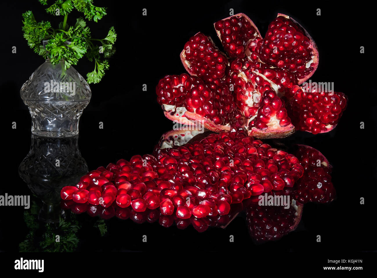 Reifer Granatapfel mit Samen spiegelt sich auf schwarze geöffnet. Kleine wider Glas Vase mit grüner Petersilie im Hintergrund. Stockfoto