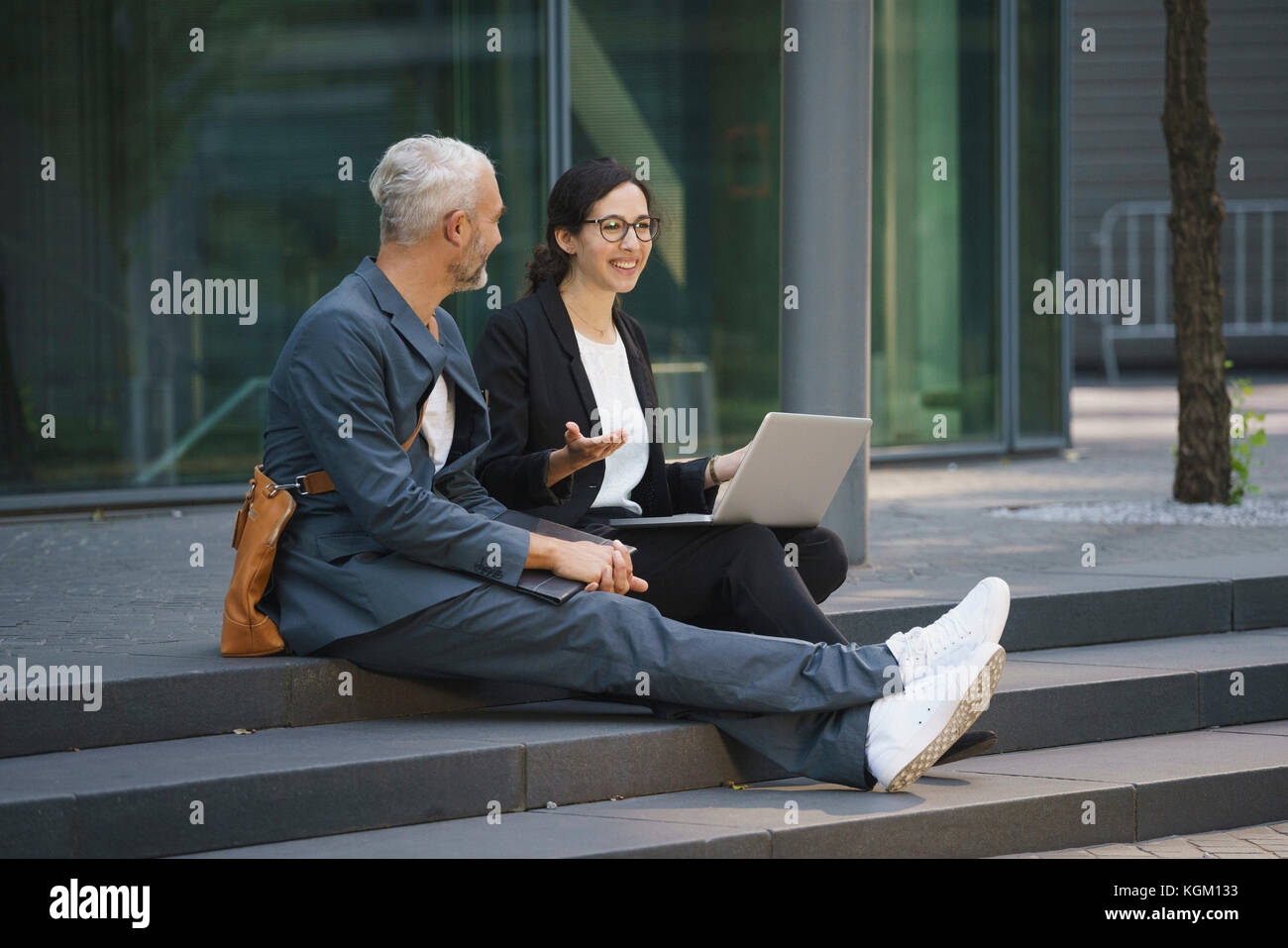 Lächelnd Kollegen mit Laptop diskutieren beim Sitzen auf Schritte in der Stadt Stockfoto
