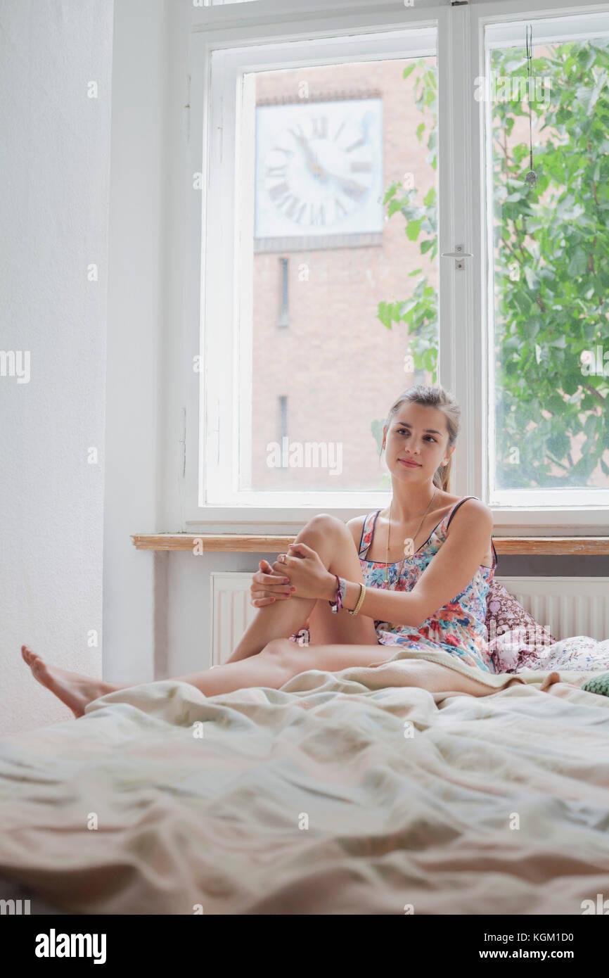 Schöne junge Frau sitzt auf dem Bett gegen Fenster zu Hause Stockfoto