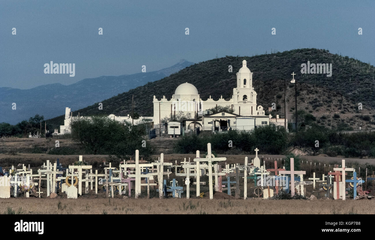 Hölzerne Kreuze markieren die Gräber der gebürtige Amerikaner ein Friedhof in der Nähe der historischen Spanischen Mission San Xavier del Bac auf der Tohono O'odham San Xavier Indian Reservation am Stadtrand von Tucson, Arizona, USA füllen. Als die "Weiße Taube der Wüste" wegen seiner hellen Kalk getünchten Fassade bekannt, die Katholische Mission war von 1783-97 durch Einheimische unter der Leitung von Franziskanerinnen gebaut. Die Mission ist eines der schönsten Beispiele spanischer Kolonialarchitektur in den Vereinigten Staaten und ist täglich geöffnet für Besucher. Stockfoto