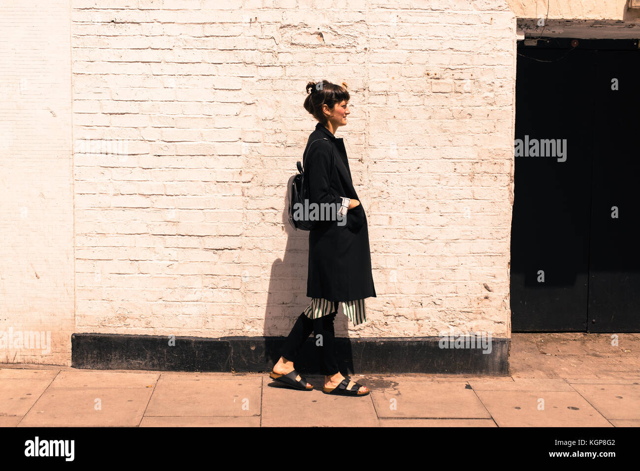 Hipster Frau in städtischen Minimal Style mit langen schwarzen Jacke, gestreiftes Kleid, schwarze Hosen und Sandalen bekleidet. Schuß auf eine Straße mit weißen Wand Stockfoto