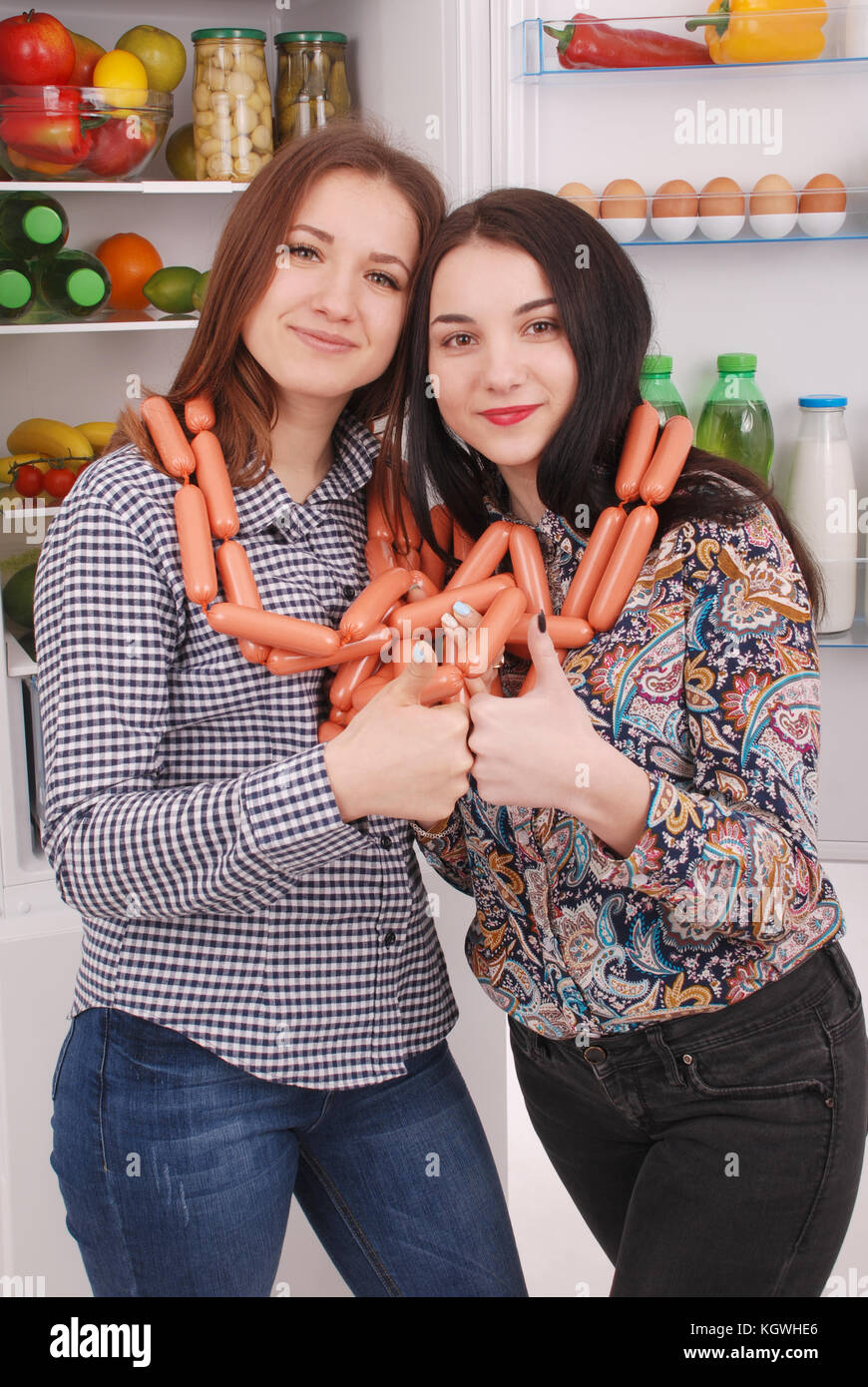 Zwei junge Mädchen hält Würstchen auf dem Kühlschrank Hintergrund. Zwei schöne junge Mädchen in der Nähe der Kühlschrank. Stockfoto