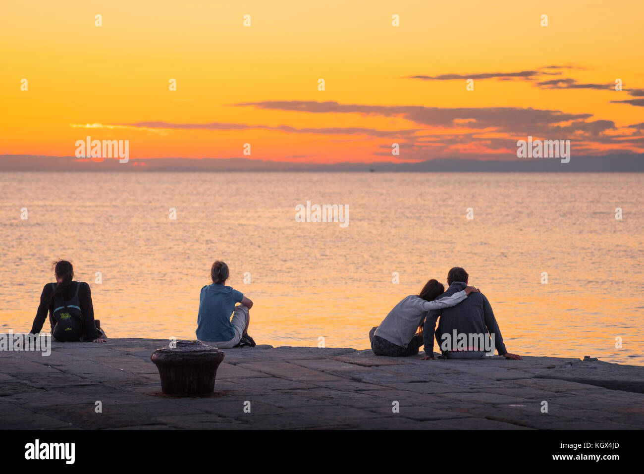 Menschen Meer romantisch, Rückansicht von jungen Menschen sitzen auf einer Hafenmauer und beobachten einen Sonnenuntergang an der Adria, Triest, Italien. Stockfoto