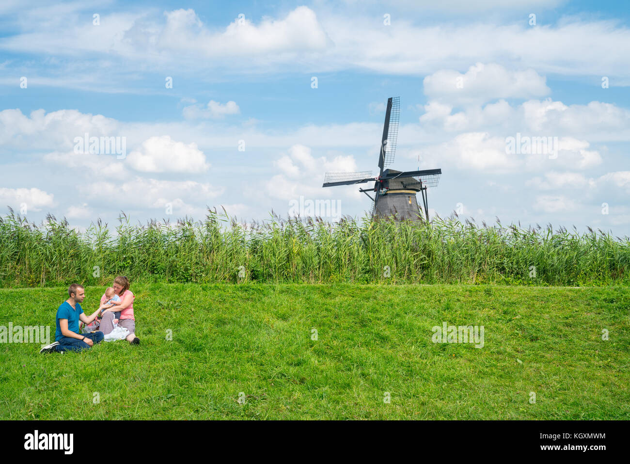 Rotterdam, Niederlande - 22 August, 2017; junges Paar mit Baby sitzen auf dem Rasen hinter dem Deich mit Windmühle in kinderdijk Bezirk beliebte touristische Destina Stockfoto