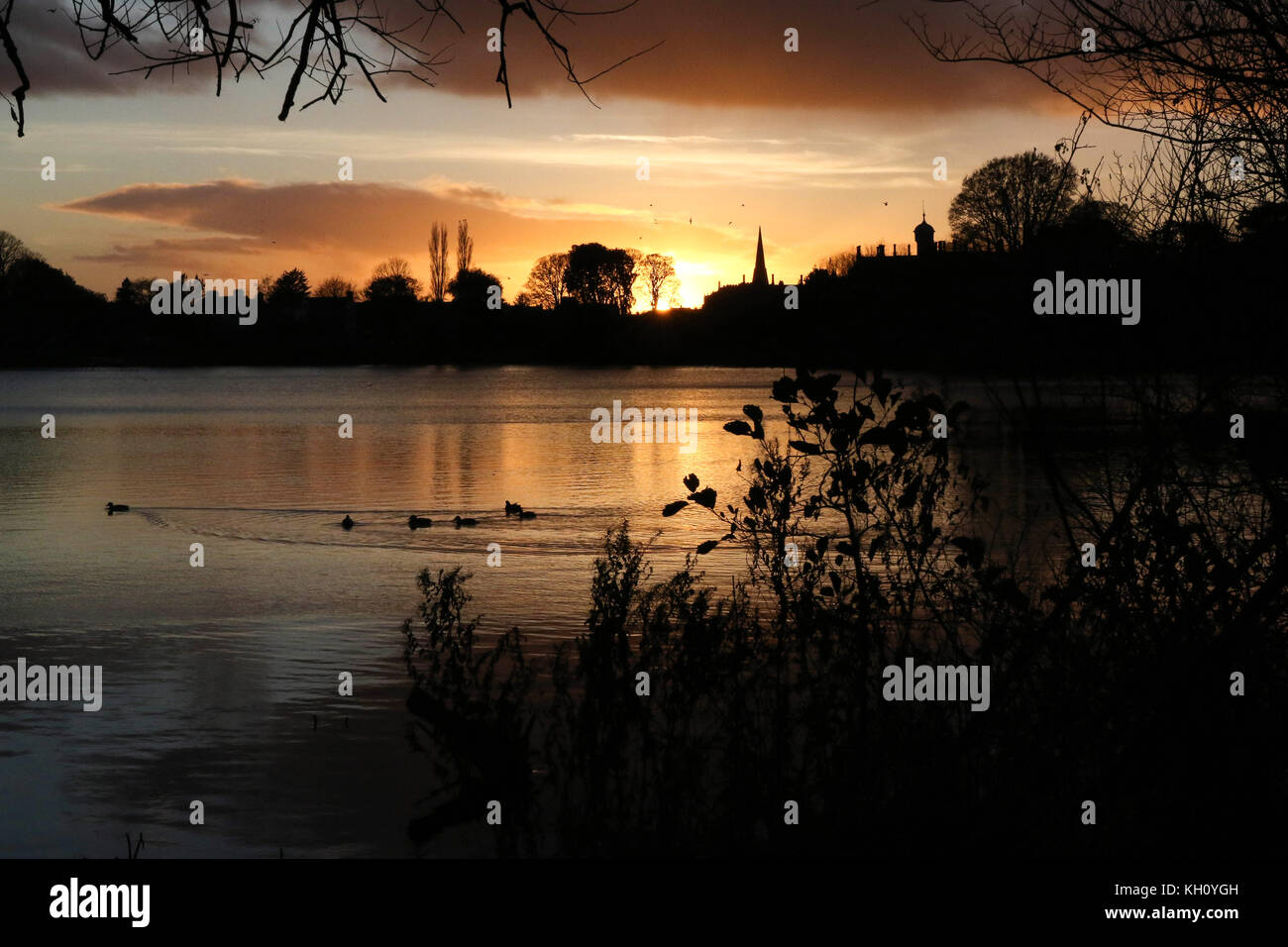 Lurgan Park See, Nordirland, Großbritannien. 12. November 2017. Ein blustery Tag mit kalten Nord-westlichen Winden und Duschen endet mit Clearing Skies und fallende Temperaturen. Frost zu rechnen ist, möglicherweise als niedrig zu-4C in ländlichen Gebieten, wie die Sonne auf der linken Seite von Shankill Pfarrkirche und Brownlow House (rechts). Quelle: David Hunter/Alamy Leben Nachrichten. Stockfoto
