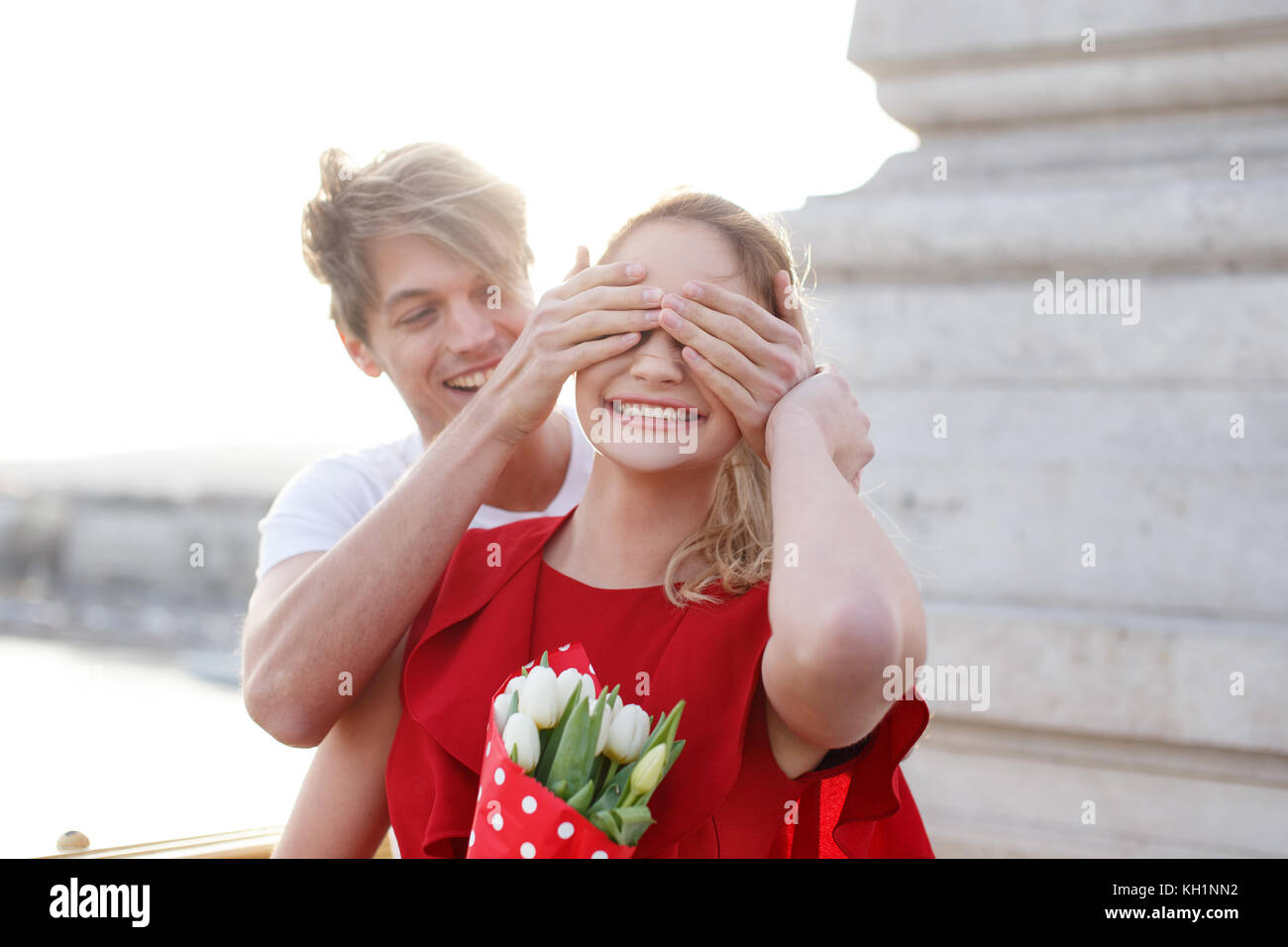 Junger Mann verstecken girlfiends Augen, Überraschung auf dating, Frau im roten Kleid mit Blumenstrauß Stockfoto
