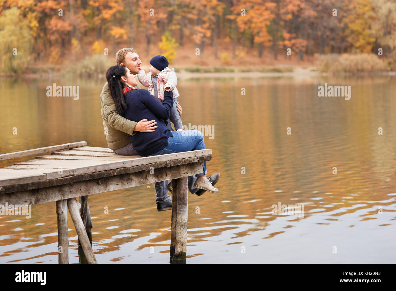 Junge Eltern sitzen auf hölzerne Brücke und mit kleinen Tochter Herbst Wald und Fluss im Hintergrund spielen. mom Holding Baby Mädchen in die Hände. Stockfoto